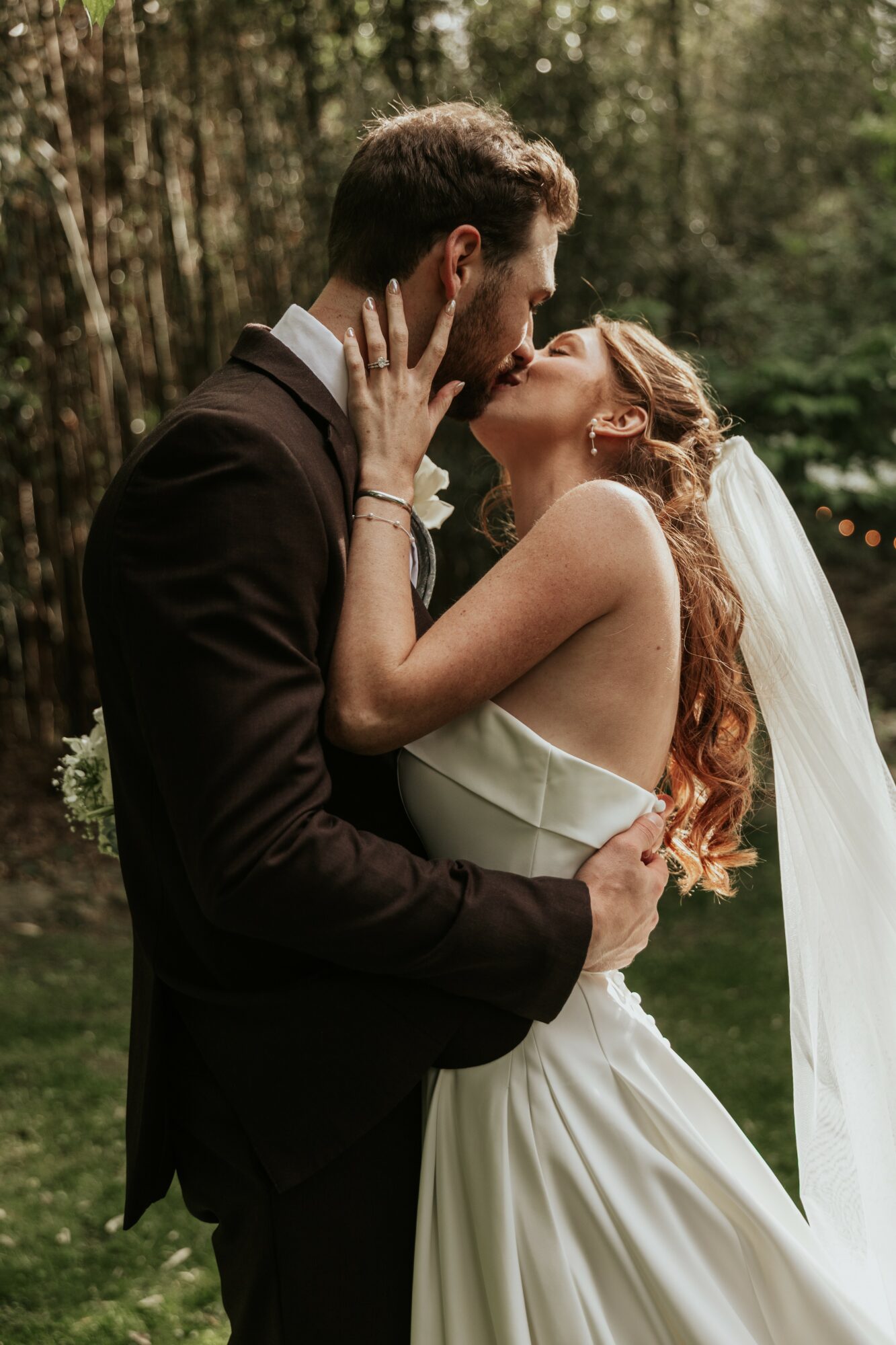 A bride and groom kiss outdoors, the bride with long red hair and a white dress, the groom in a dark suit.