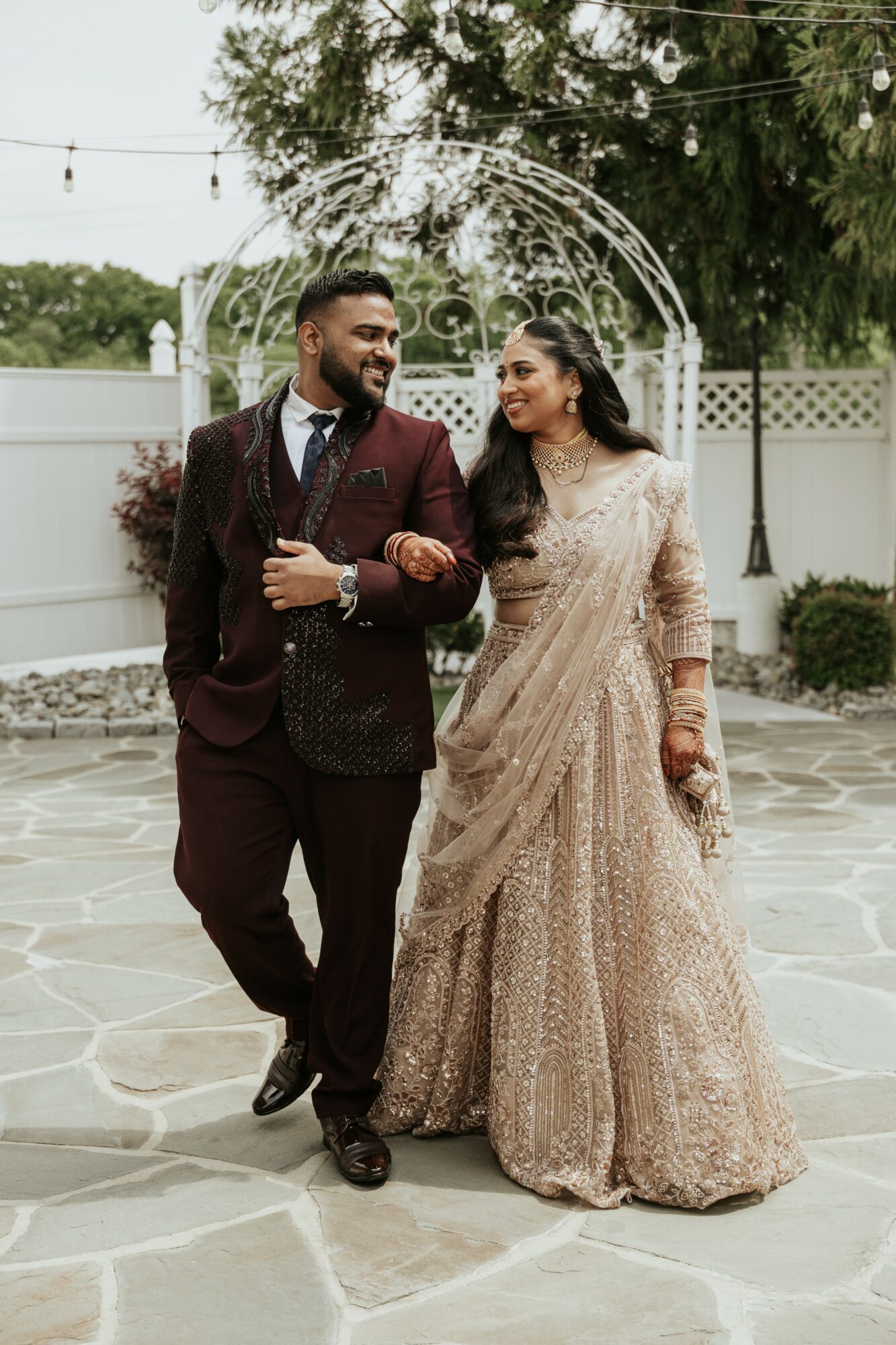 A couple dressed in formal attire walking outdoors on a stone pathway, smiling and holding hands, with trees and a white fence in the background.