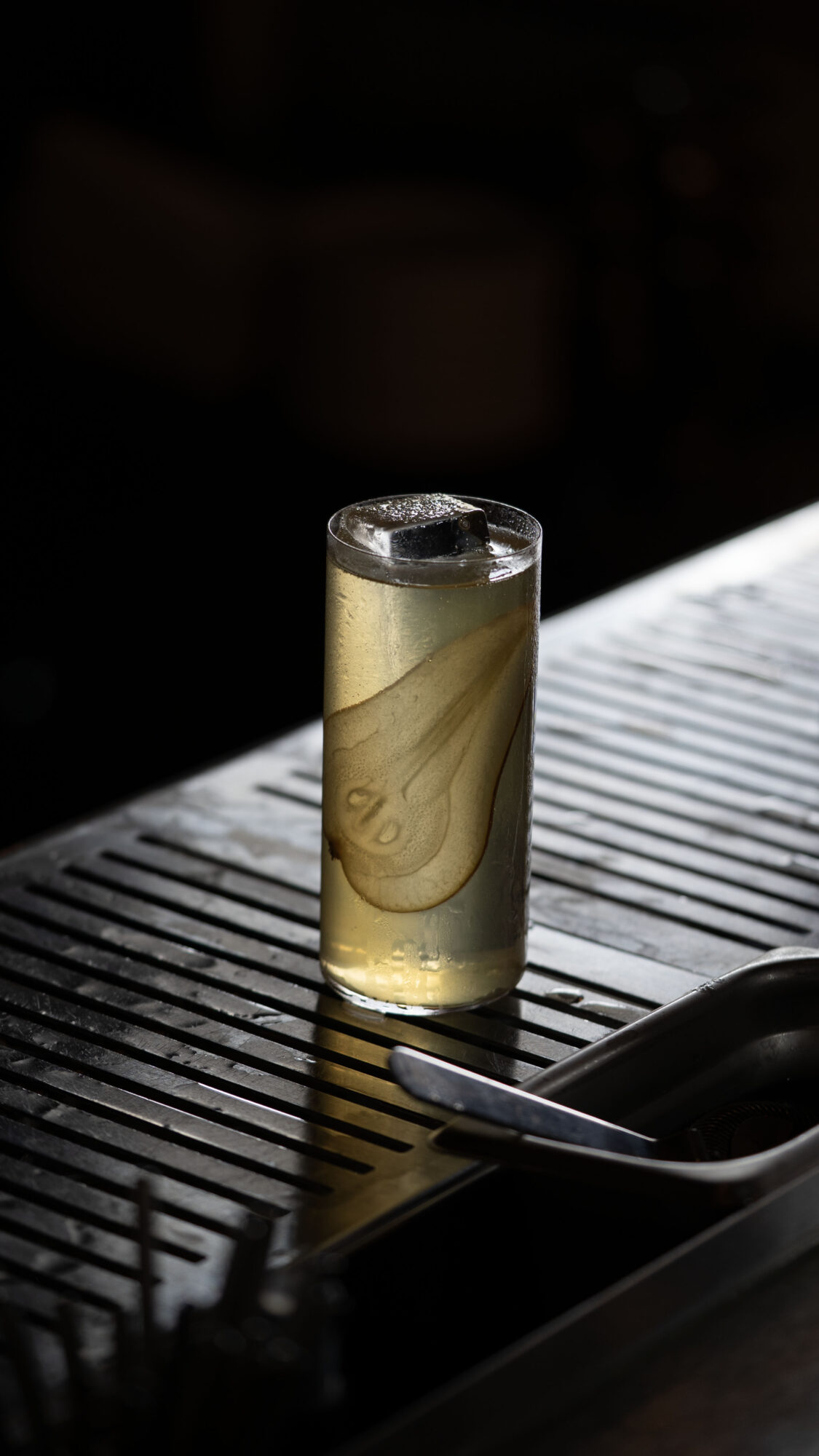 A tall glass with a beverage and ice cube on a metal surface, with a spoon nearby, in a dim setting.