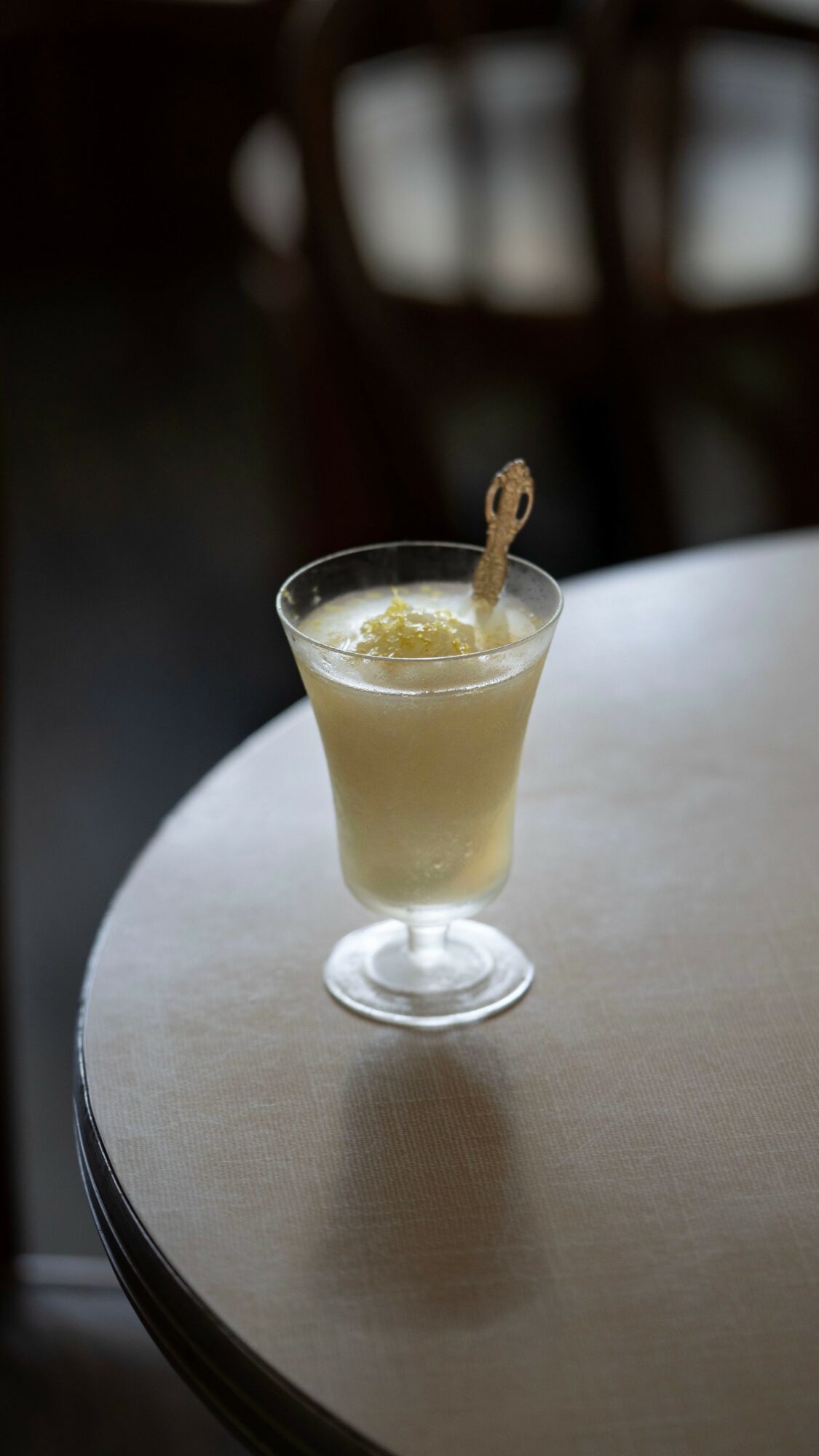 A cocktail glass with a light-colored drink and a garnish on a round table, background blurred.
