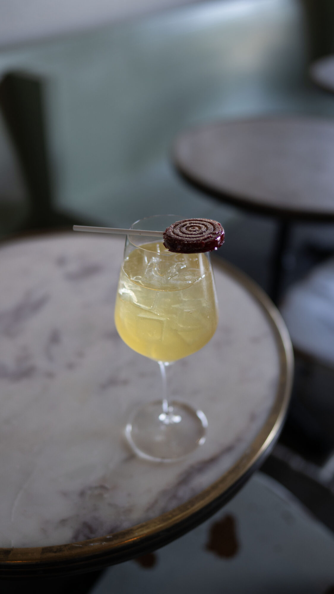 Yellow cocktail with ice in a glass, garnished with a spiral chocolate piece and a straw, on a marble table.