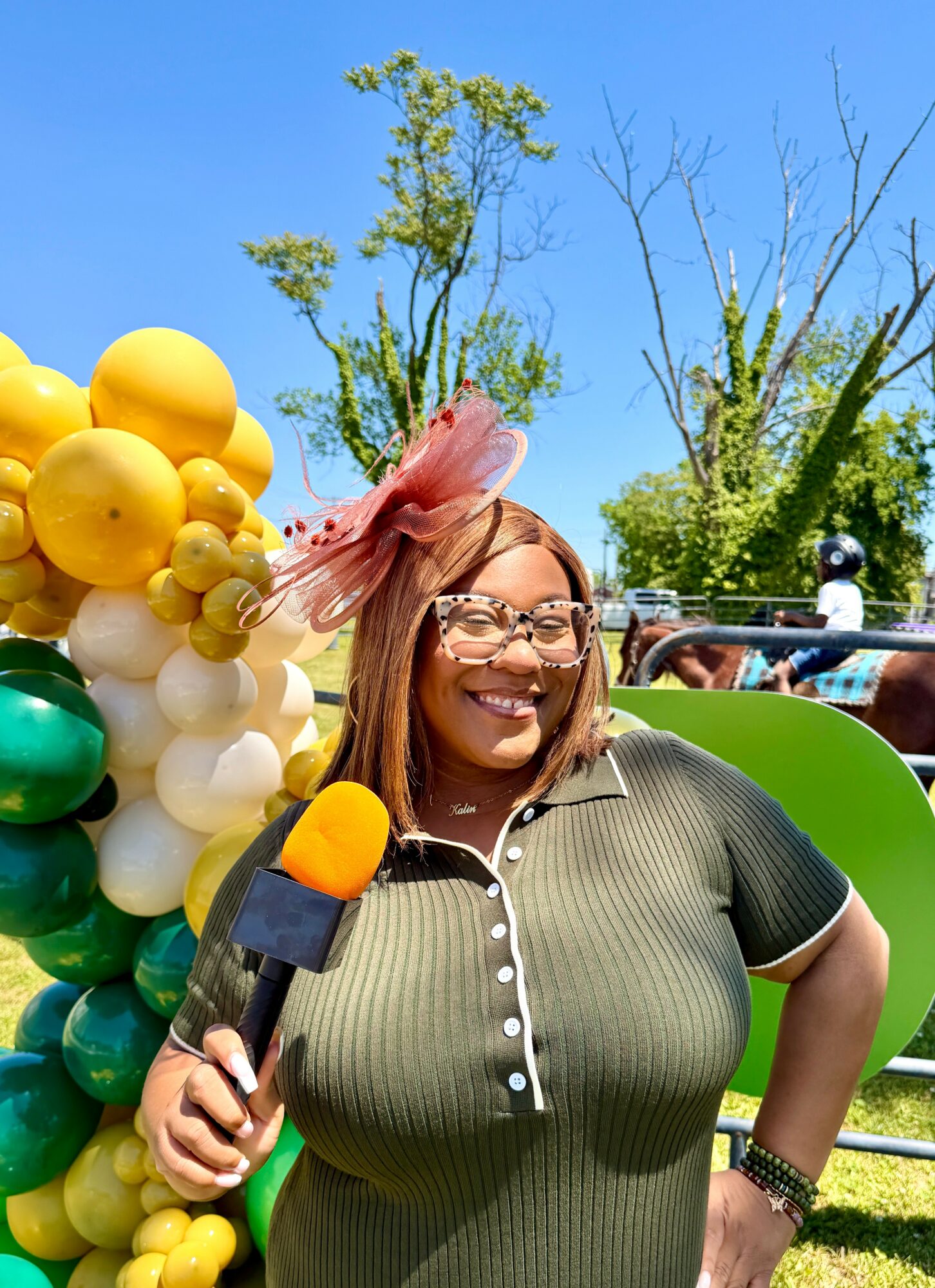 Smiling woman with glasses holding a microphone outdoors, decorated with colorful balloons and trees in background.