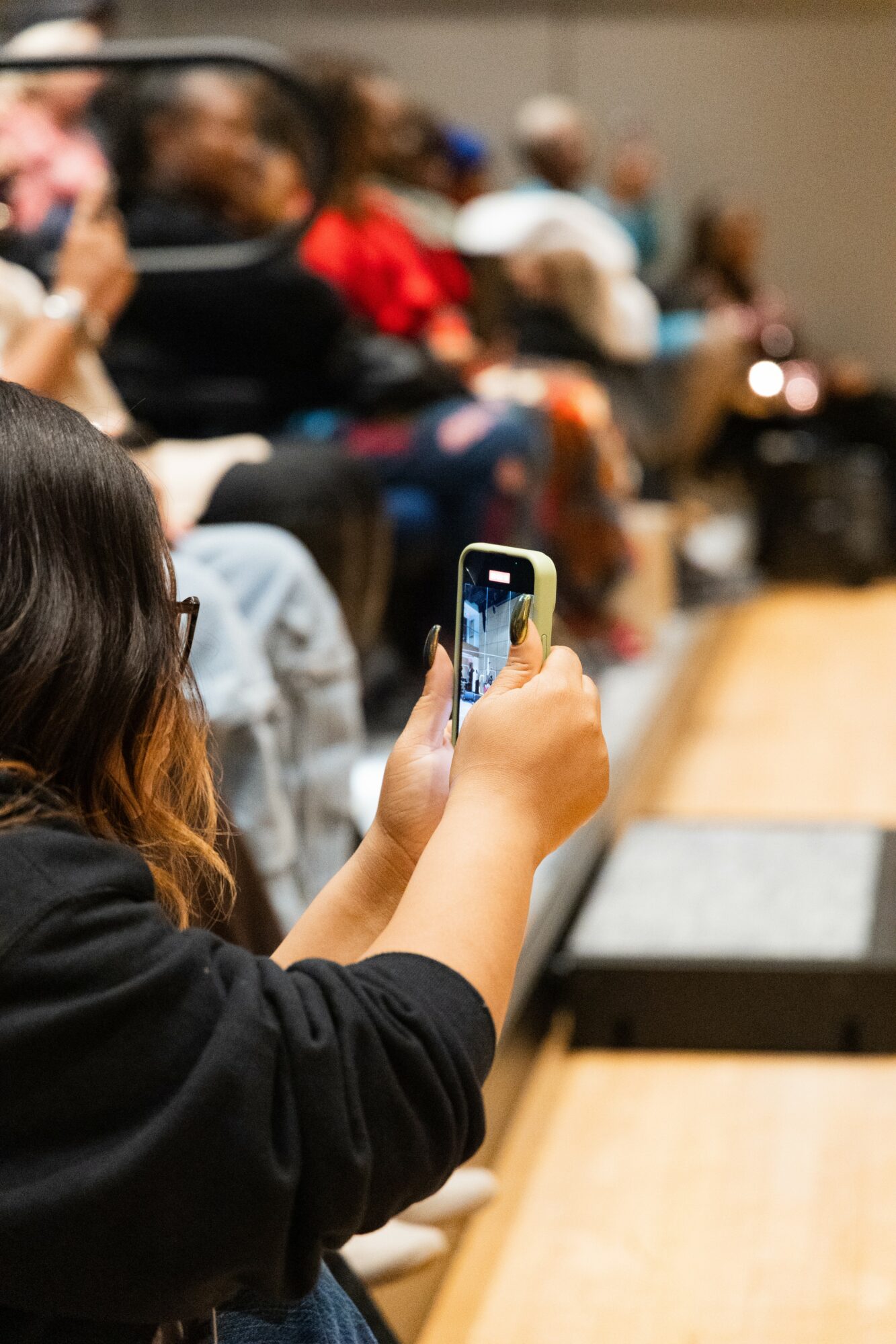 Person taking a photo with a smartphone in a room with seated audience members, some wearing red clothing.