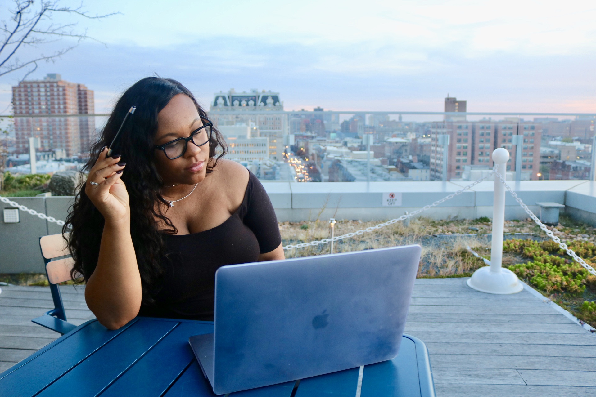 Woman with glasses working on a laptop on a rooftop with city skyline in background.
