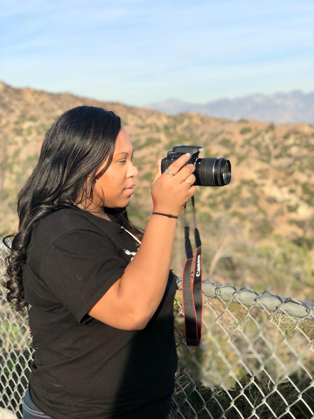 Woman with long dark hair holding a camera outdoors, with a landscape of hills and sky in the background.