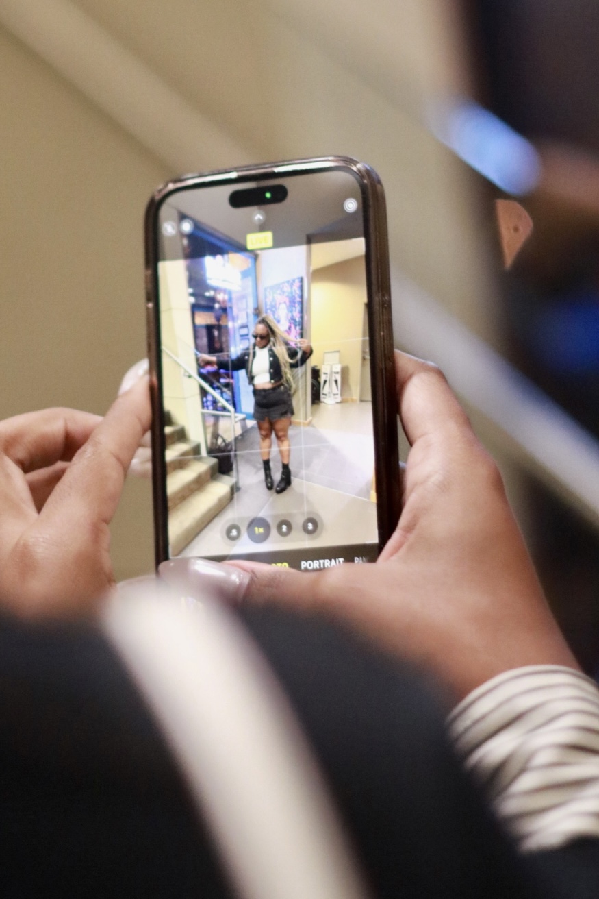 Person taking a photo of a woman in a store using a smartphone, with shelves and bright lighting visible.