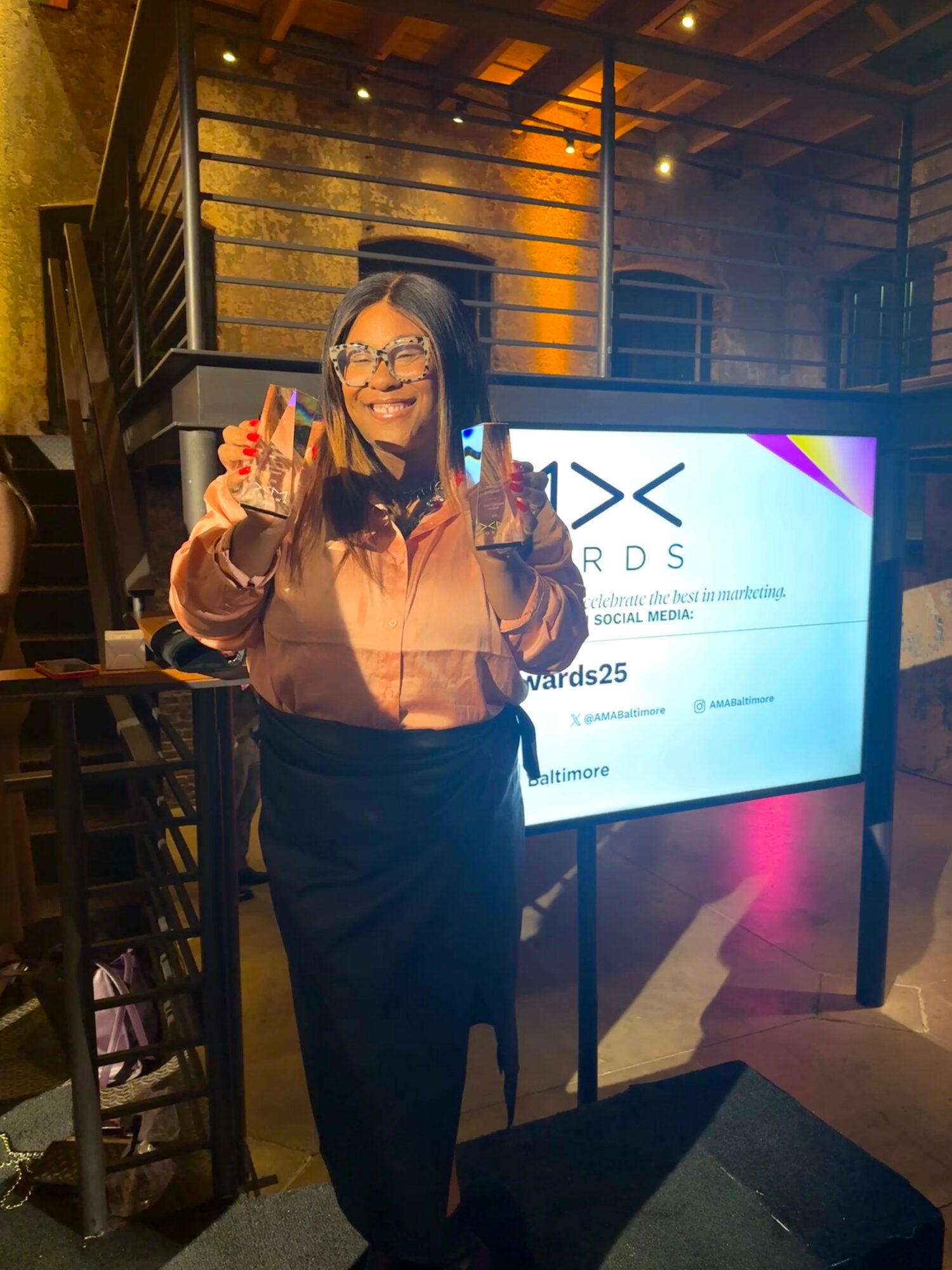 Woman with glasses and long hair, smiling, holding up two peace signs, standing indoors near a digital display.