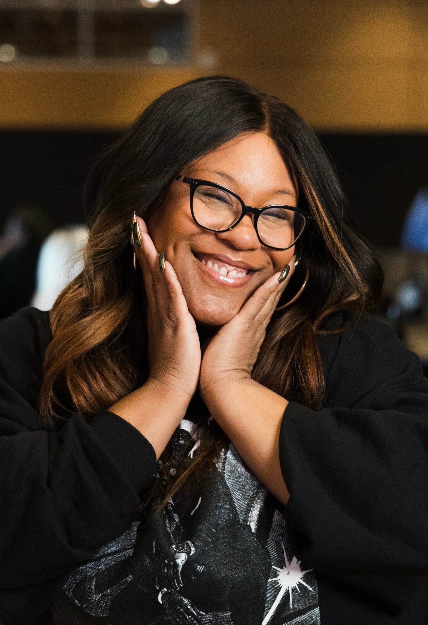Smiling woman with glasses and long hair, hands on face, in a dark top, in an indoor setting.
