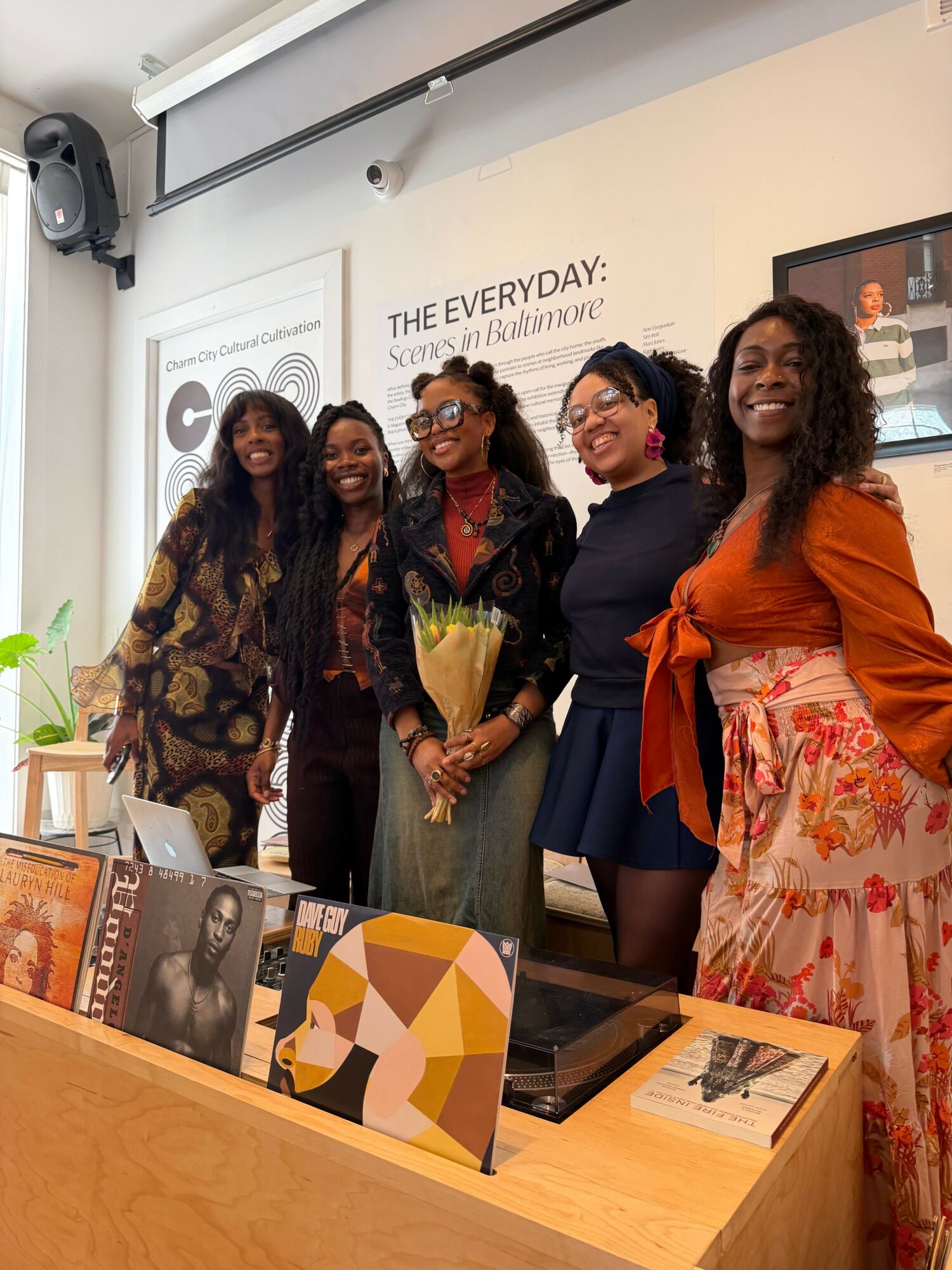 Four women standing together in a room, holding a bouquet of flowers, smiling, with a white wall and a poster behind them.