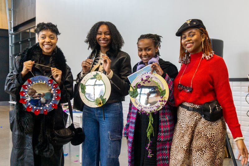 Four women smiling and holding decorative circular art pieces, standing indoors against a plain wall.