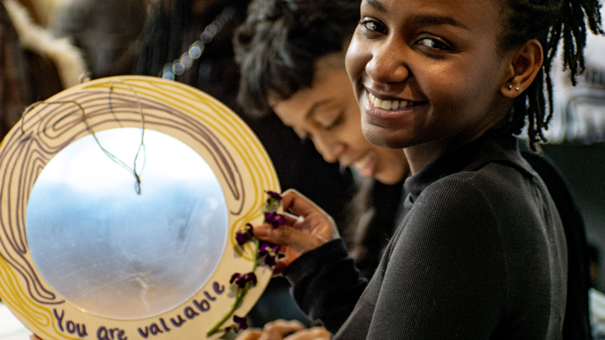 Smiling woman holding a decorated mirror with a message, surrounded by people, in a group setting.