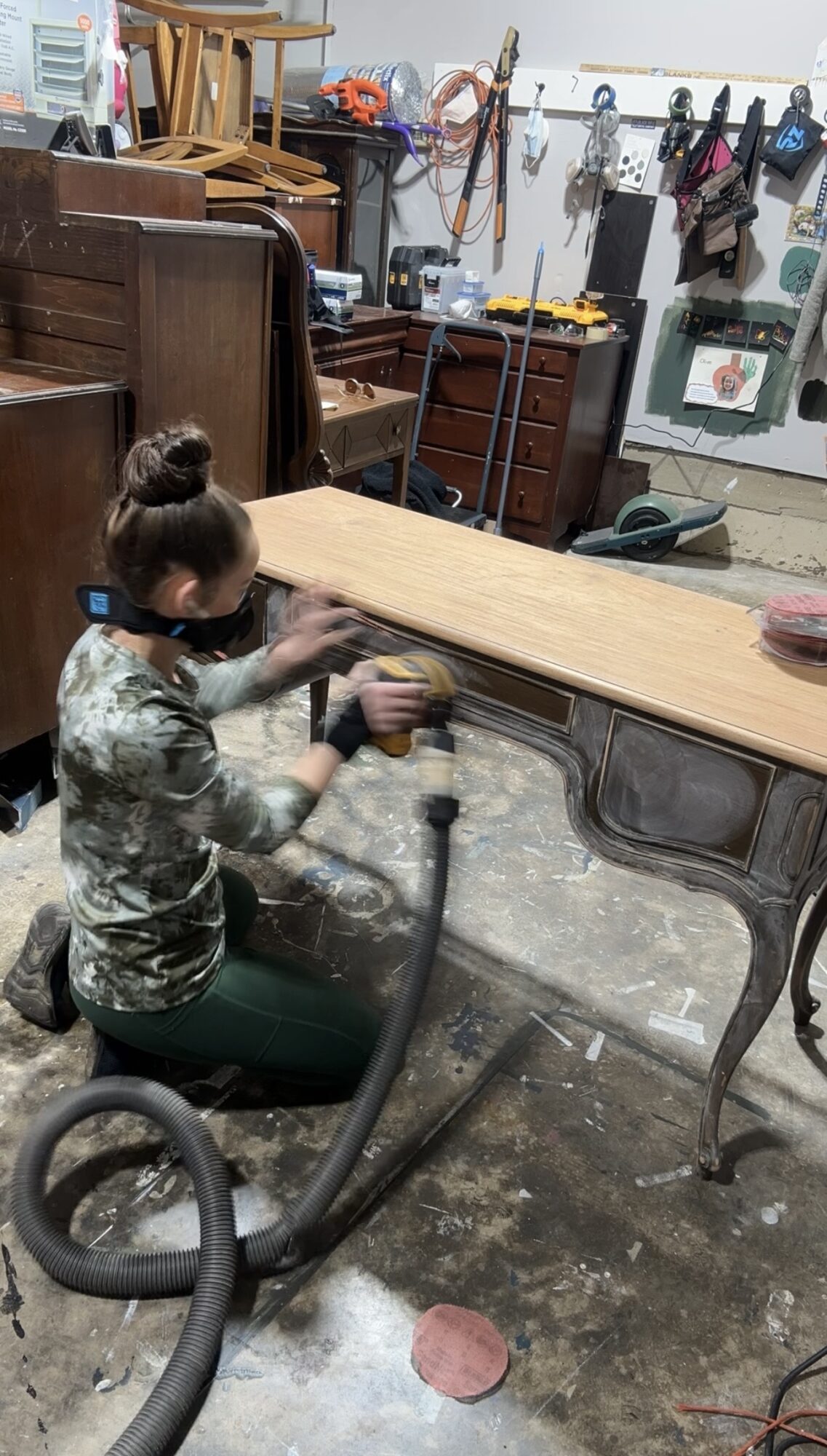 Person cleaning a wooden table in a cluttered room with furniture, tools, and hanging items, using a vacuum cleaner.