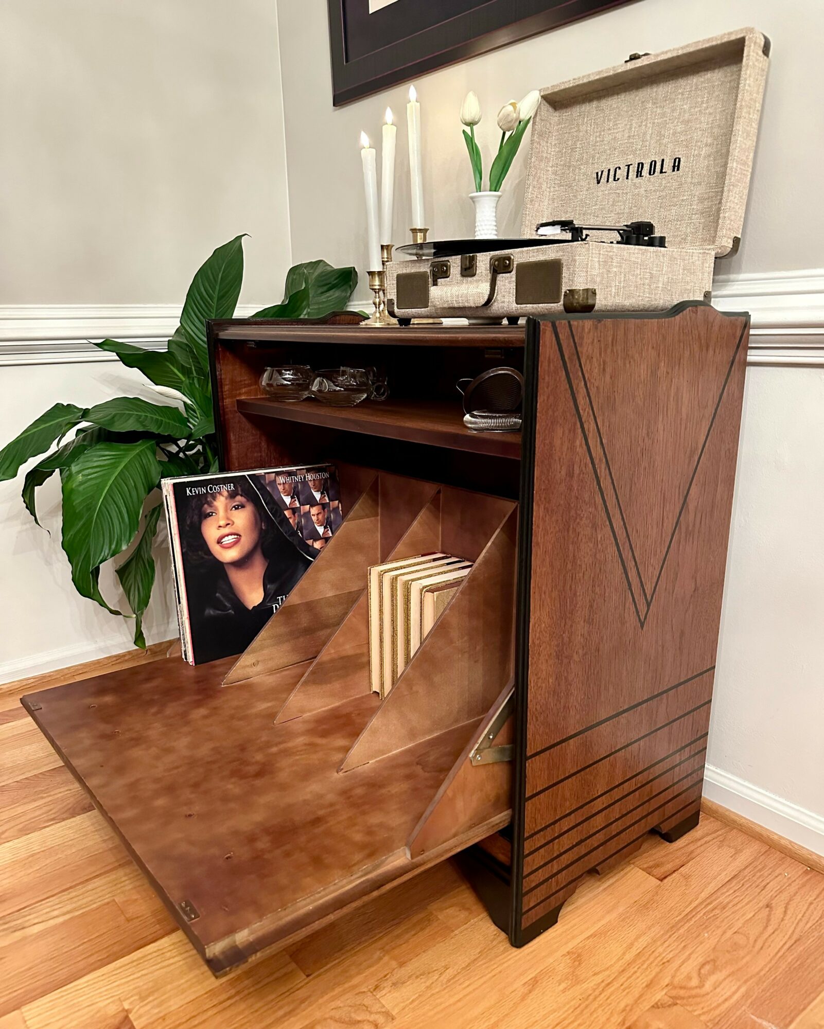 Wooden record player stand with a record player on top, a large green plant, and a framed photo leaning against the stand.