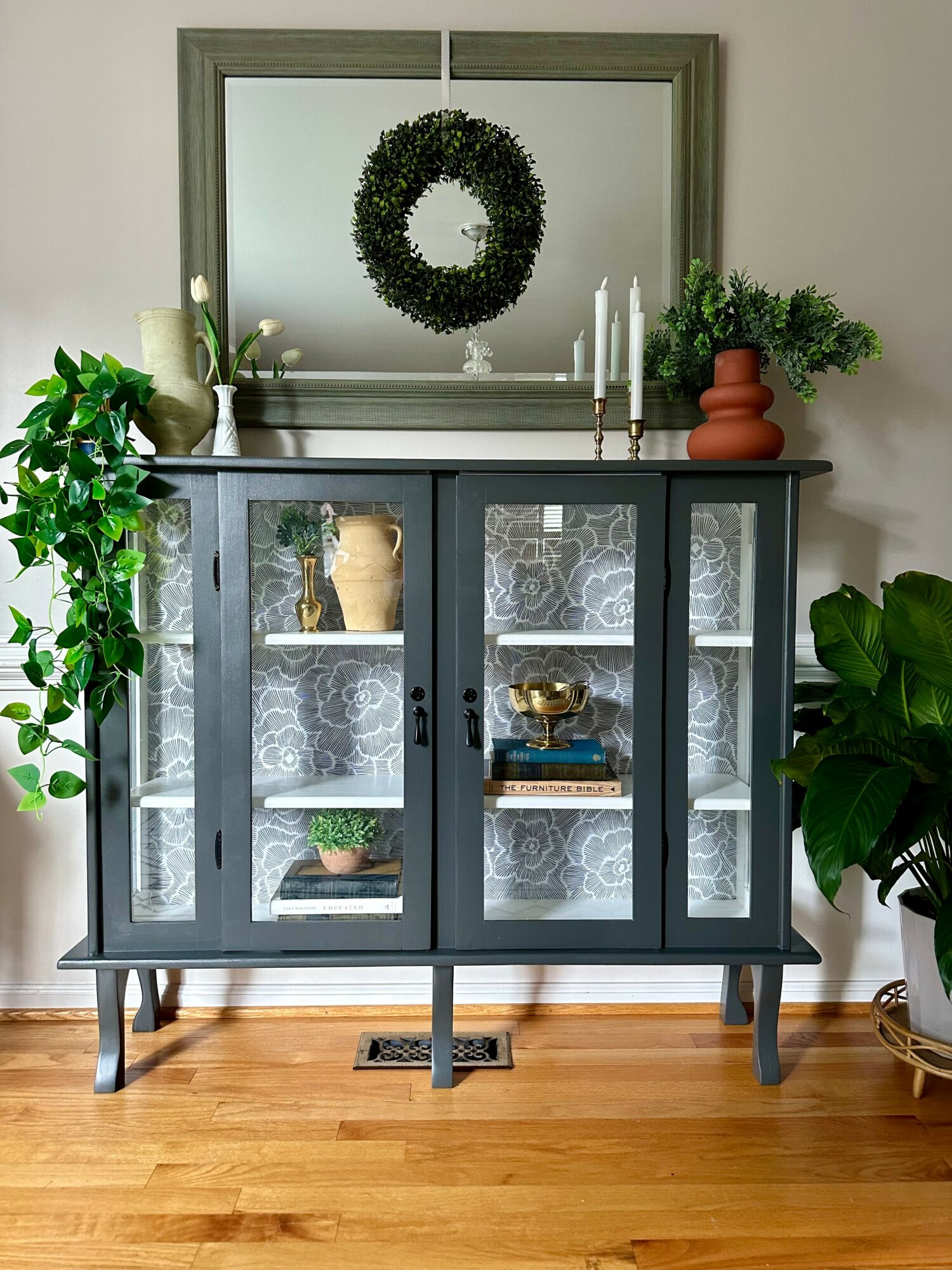 Gray cabinet with glass doors, decorative lace panels, and items inside, flanked by potted plants, with a mirror and wreath above.