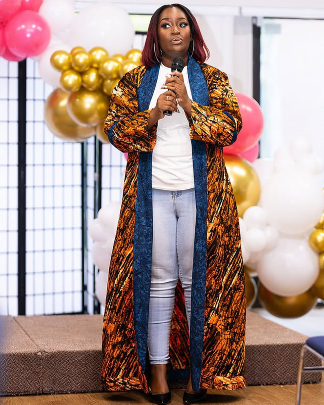 Woman standing on stage with balloons, holding a microphone, wearing a long patterned coat and jeans.