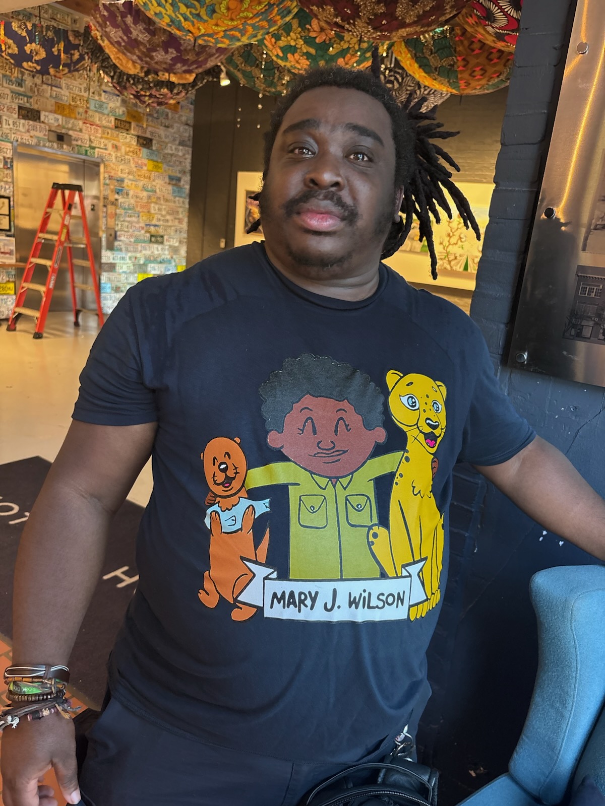 Person with dreadlocks wearing a cartoon character T-shirt, standing indoors with colorful decorations in the background.