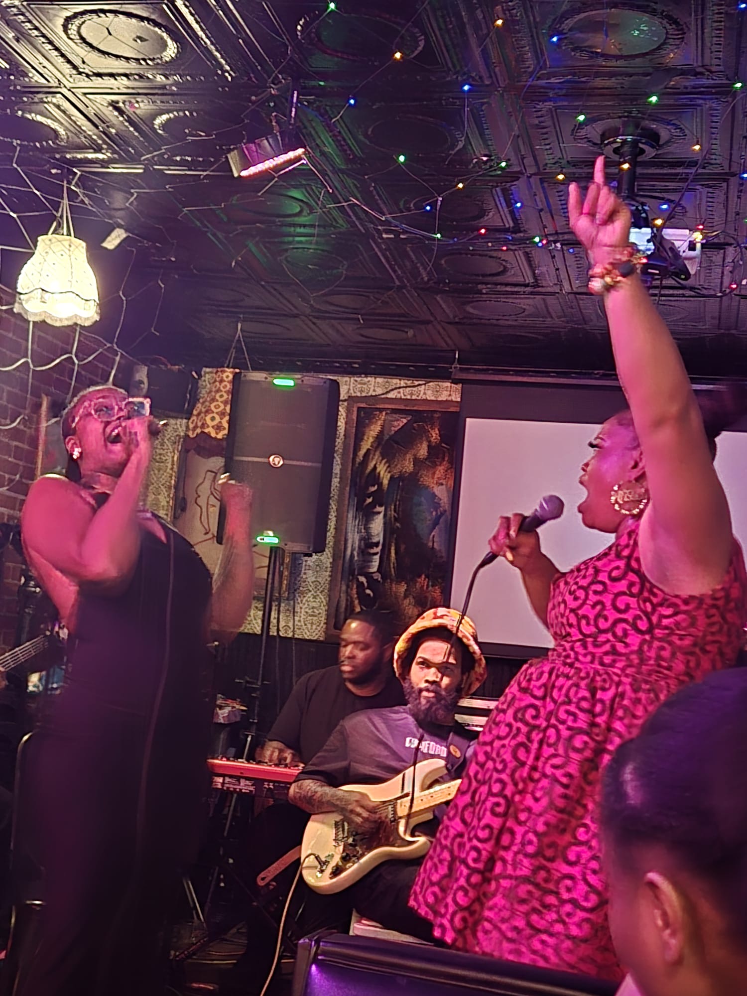 Two women singing and performing on stage with musicians, one holding a microphone and the other raising her hand, in a lively setting.