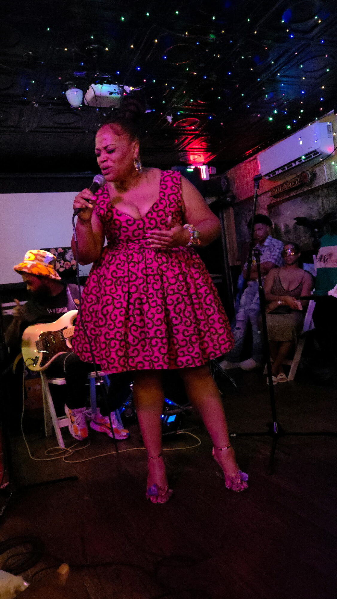 Woman in pink dress singing or speaking on stage with a microphone, surrounded by people and colorful lights.