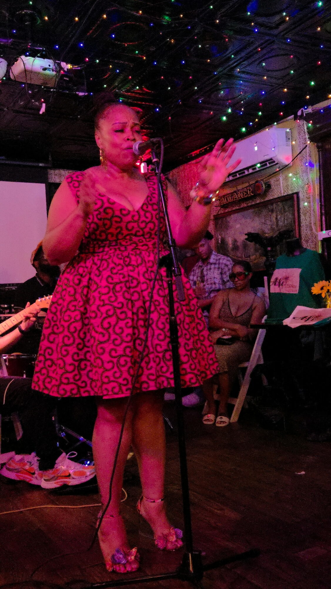 Woman in pink dress speaking into microphone at a party with colorful ceiling lights and audience in background.
