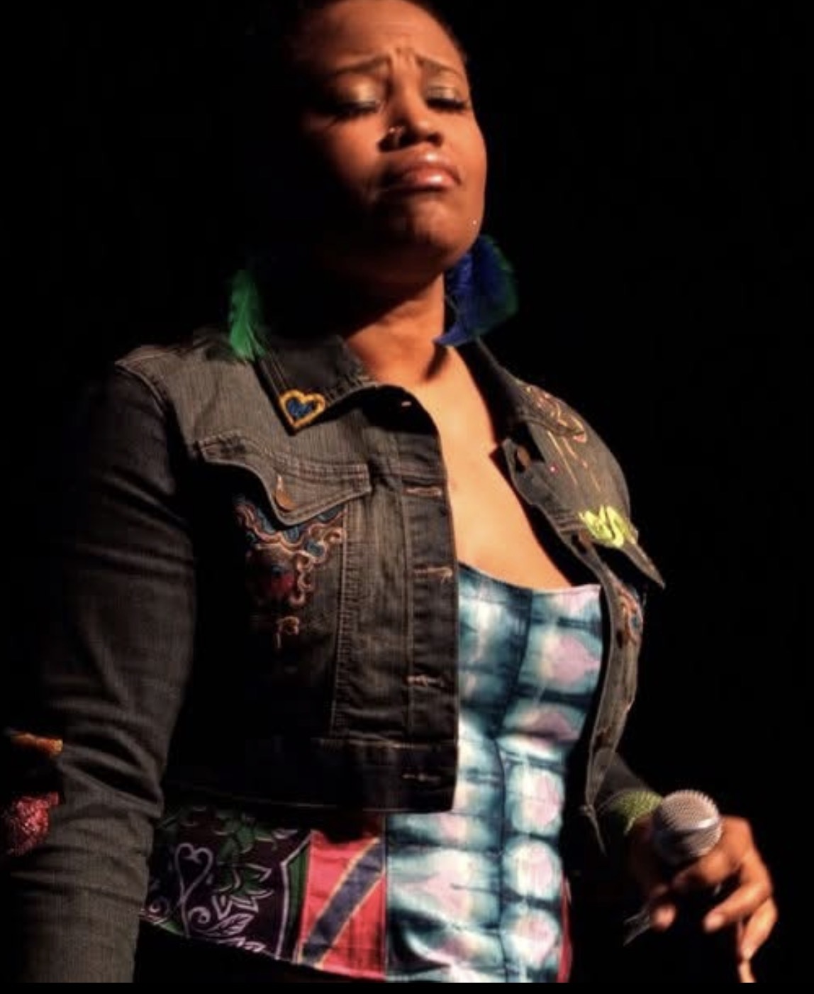 Person with closed eyes holding a microphone, wearing a denim jacket and colorful dress, on a dark stage.
