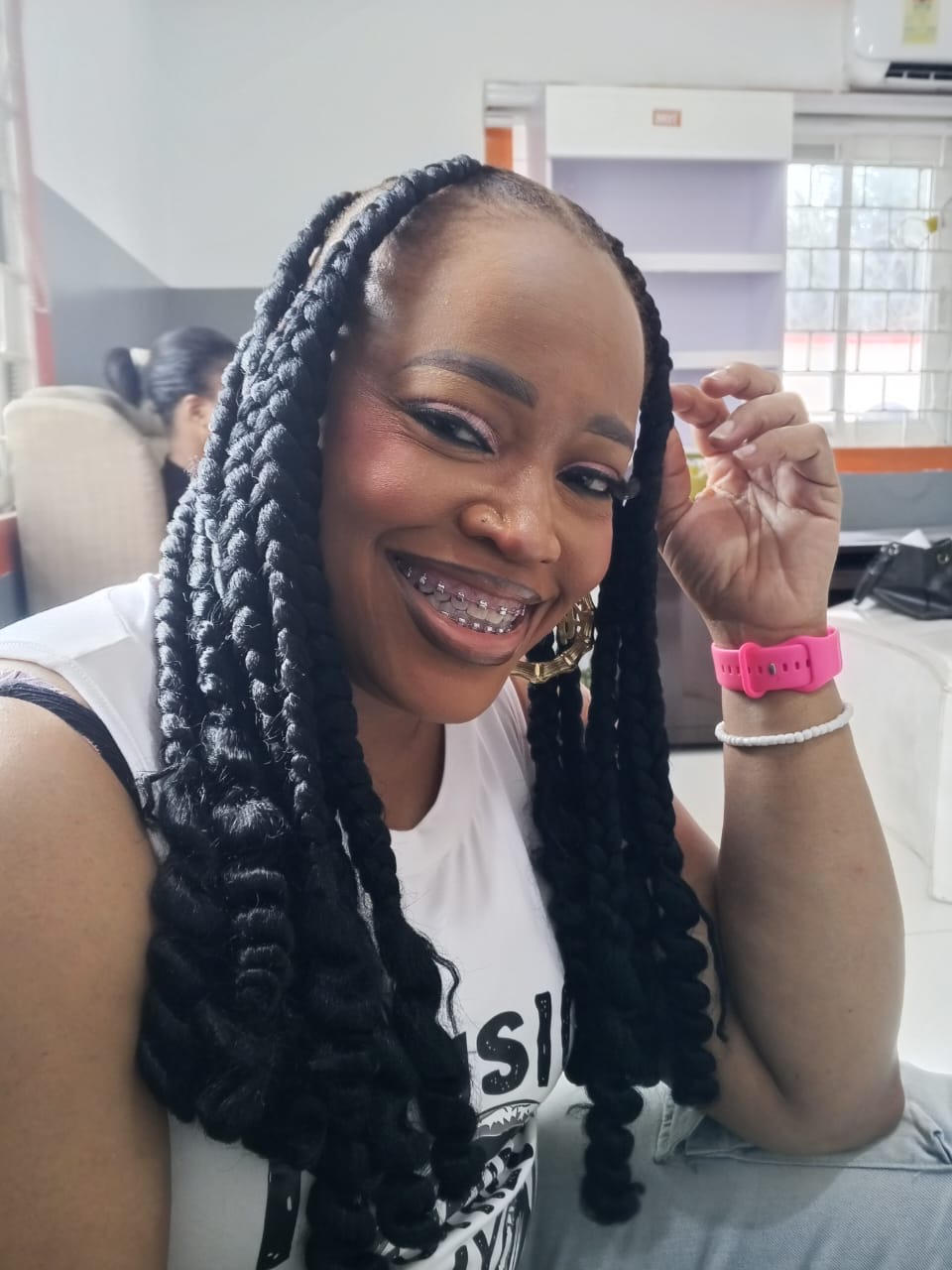 Smiling woman with long braided hair, braces, and a pink watch, sitting indoors with a bright background.