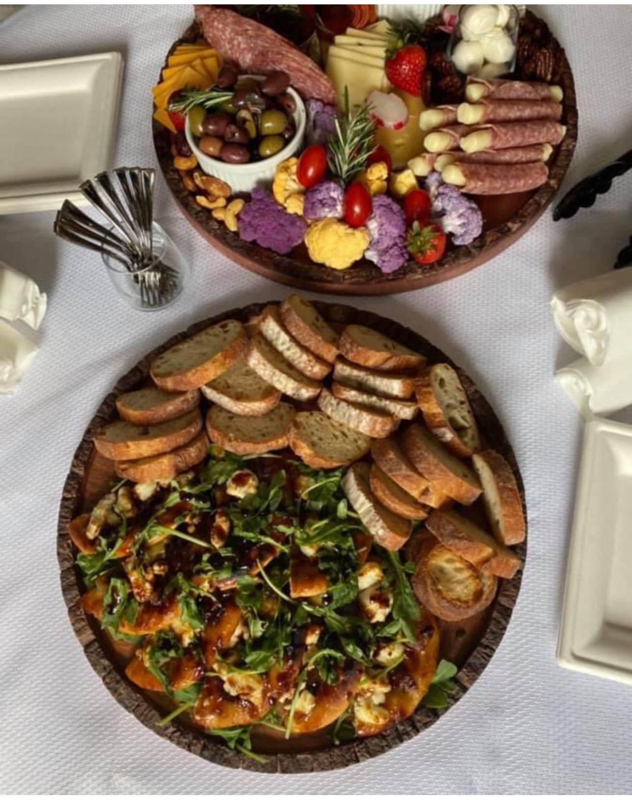 Two large platters of assorted appetizers and finger foods on a table with white tablecloth, surrounded by plates and utensils.