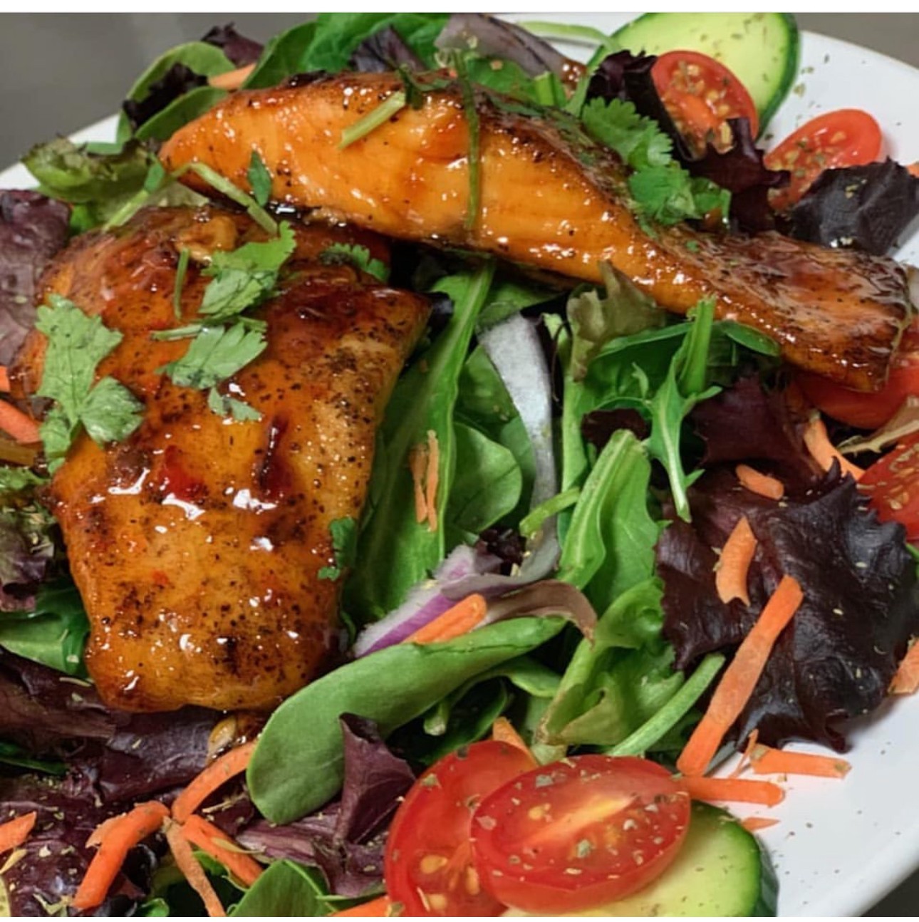 Salad with grilled fish, cherry tomatoes, cucumber, carrots, and mixed greens, garnished with cilantro.