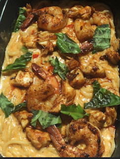 Dish with cooked shrimp, chicken pieces, pasta, and green leafy vegetables in a creamy sauce.