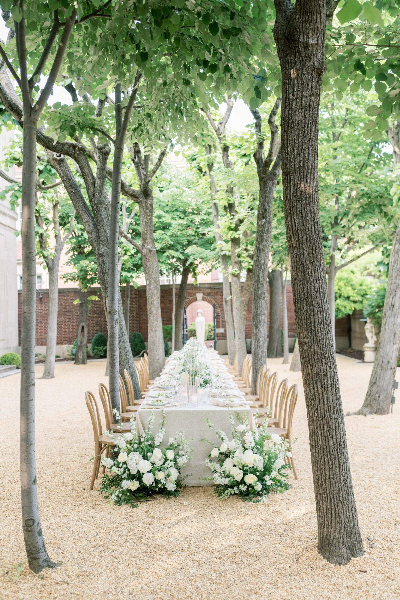 Long table set outdoors with white flowers and chairs, surrounded by trees and a brick wall in the background.