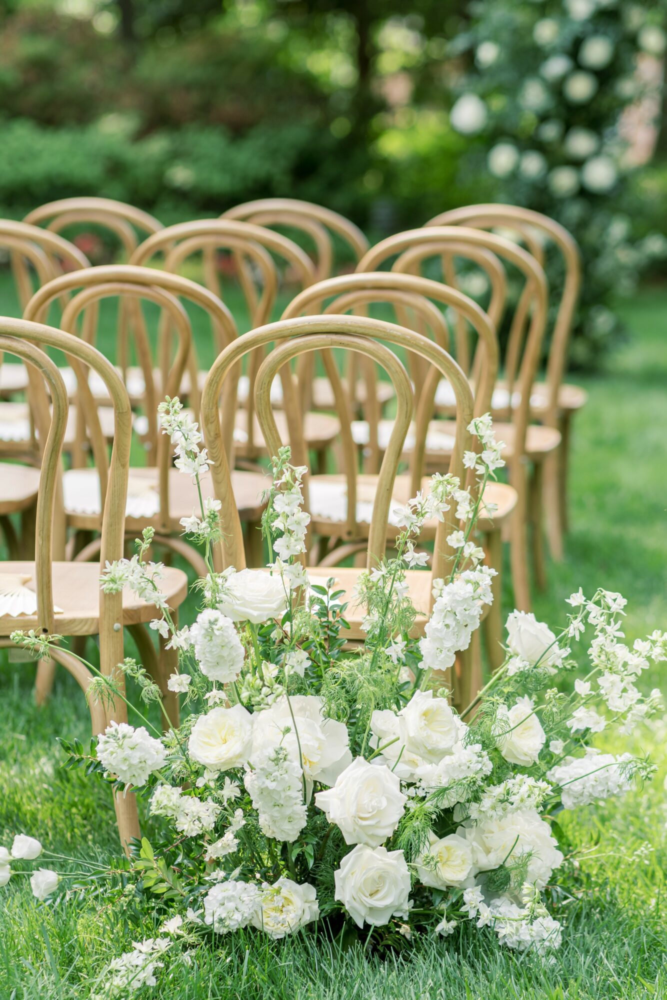 Rows of wooden chairs outdoors with white floral arrangements on green grass, garden background, sunny day.