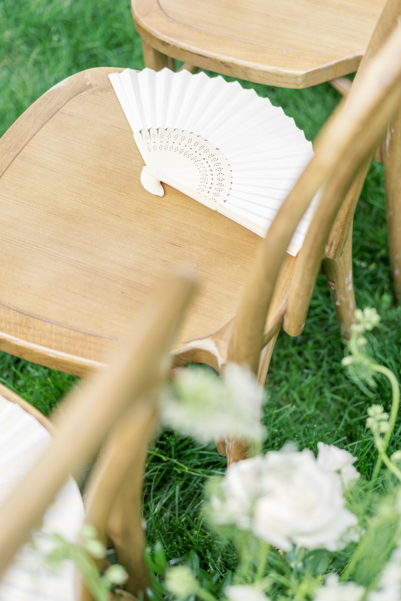Wooden chair with a white folding fan resting on the seat, outdoors on grass with white flowers in foreground.