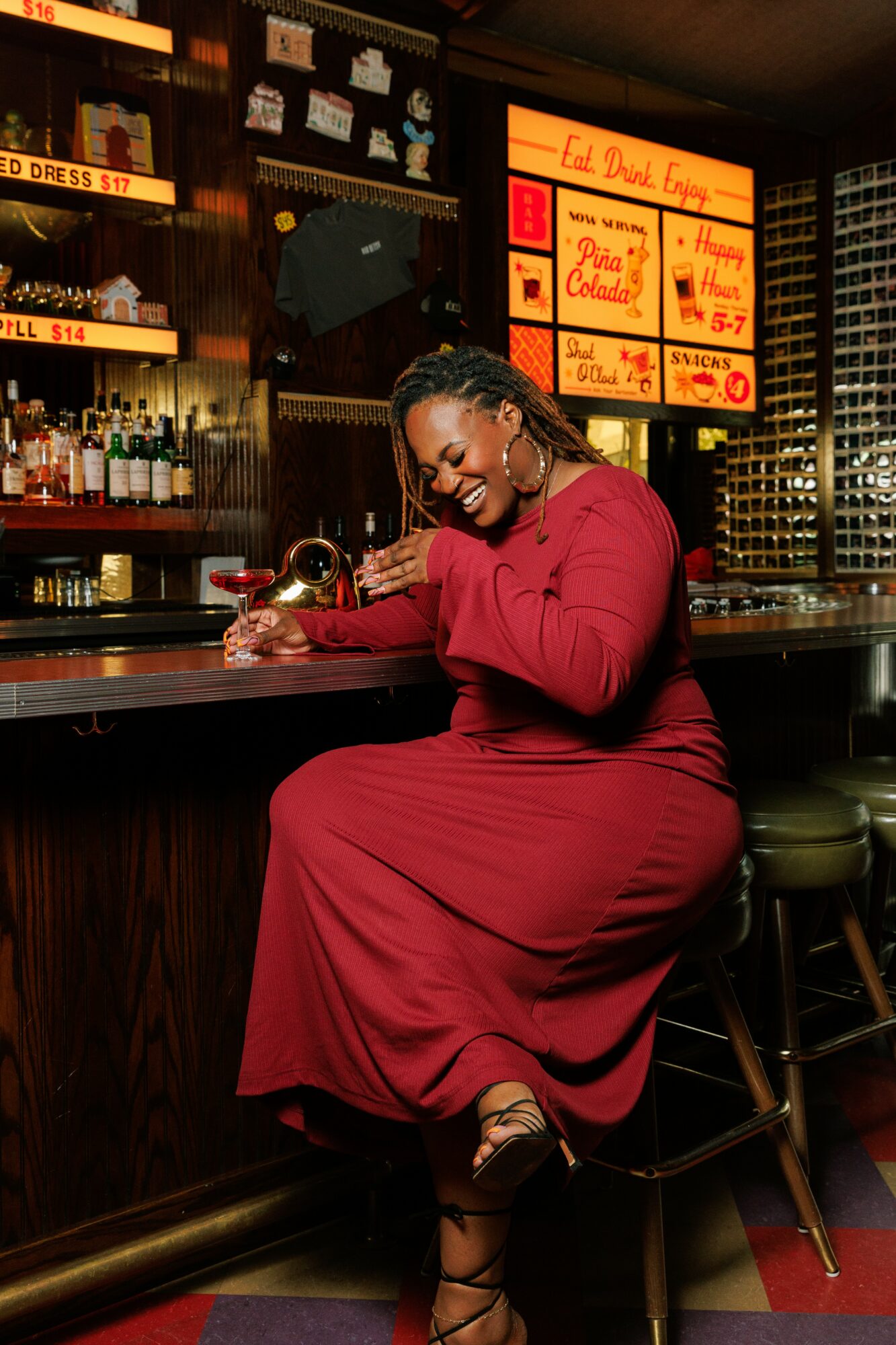 Woman in red dress sitting at bar, smiling, with bar and illuminated menu in background.