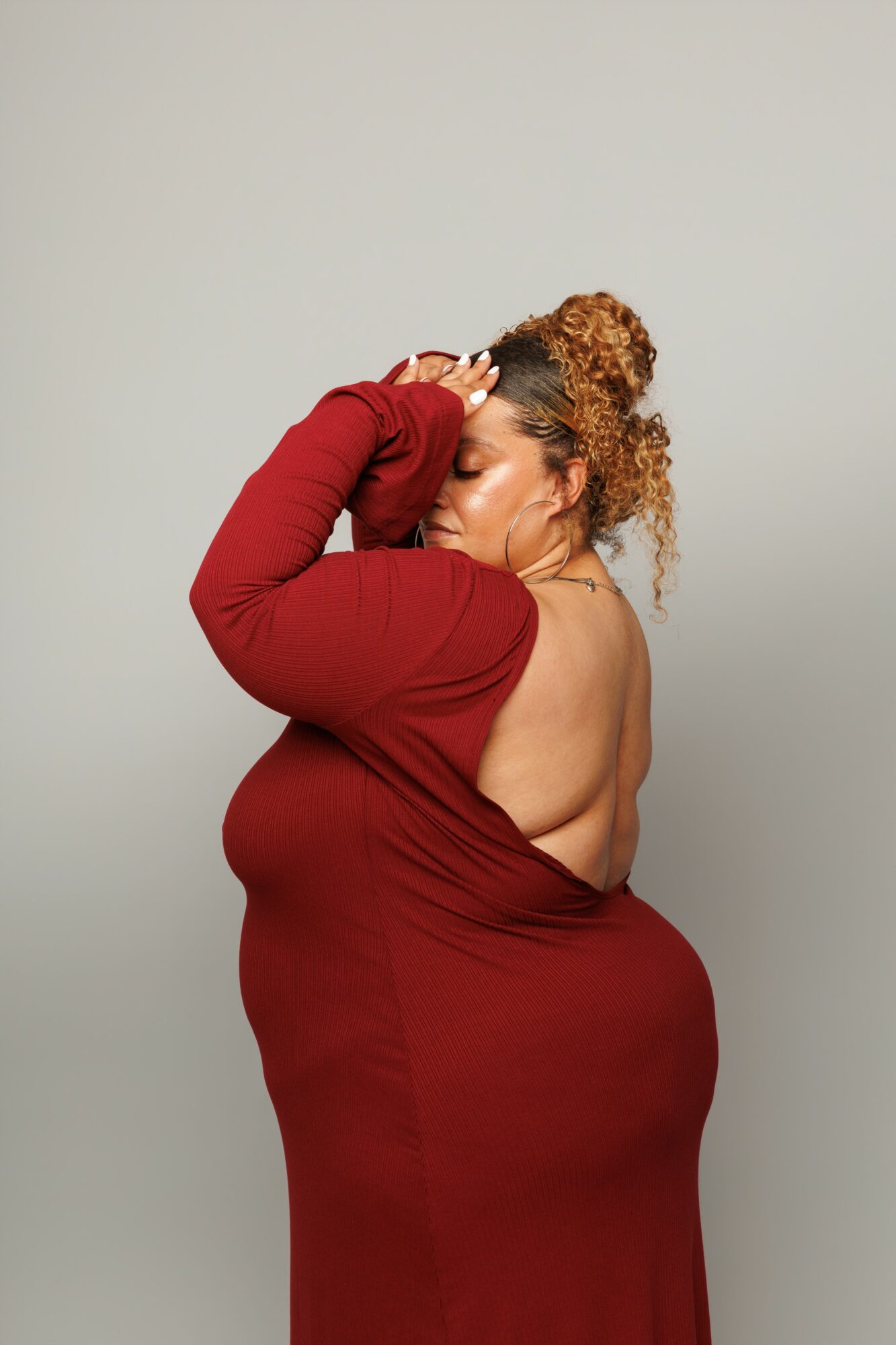 Woman in a red dress with an open back, posing with her eyes closed and hand on her head.