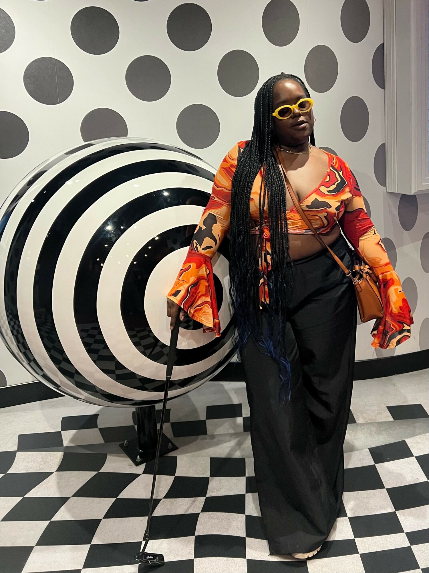 Woman with long braids, yellow sunglasses, orange patterned top, black pants, standing next to a black and white spiral sculpture in a polka dot room.