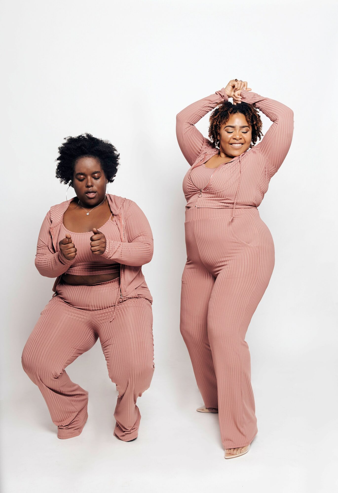 Two women in matching pink outfits pose against a white background, one with hands on head and the other with hands in front.