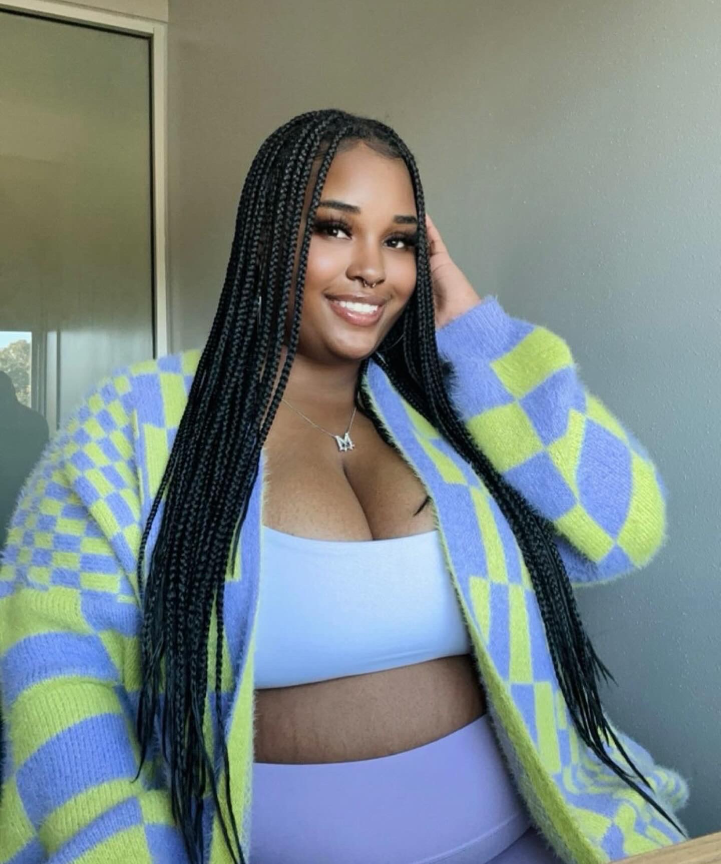 Smiling woman with long braided hair wearing a blue and green checkered cardigan and white crop top, indoors.