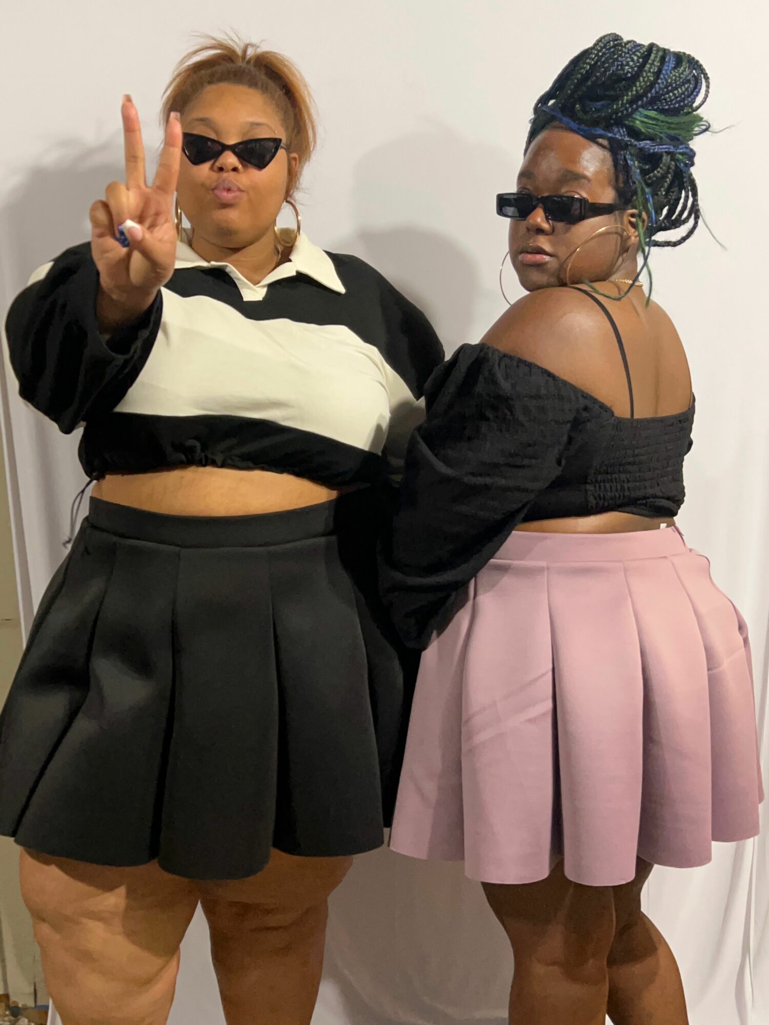 Two women posing, one making a peace sign, both wearing sunglasses and stylish outfits, standing against a plain background.