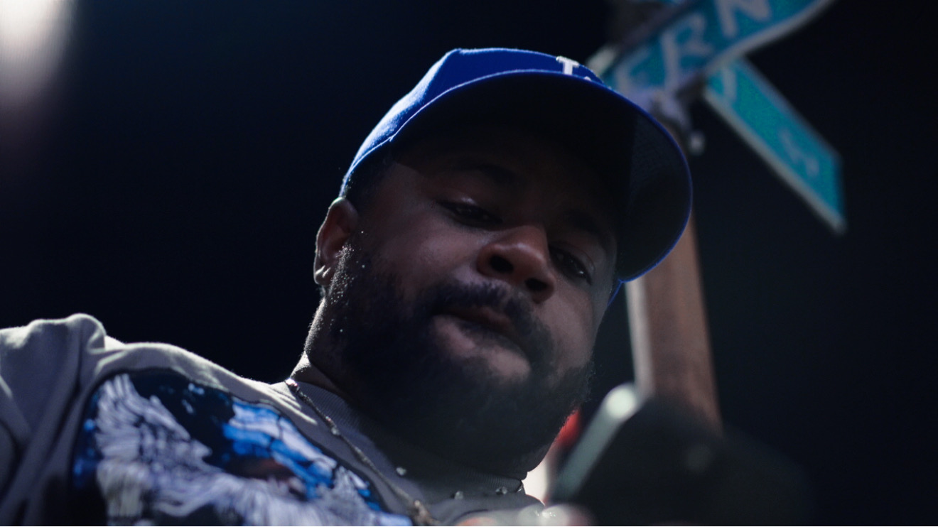 Close-up of a man wearing a blue cap, looking down, with a dark background and a street sign partially visible.
