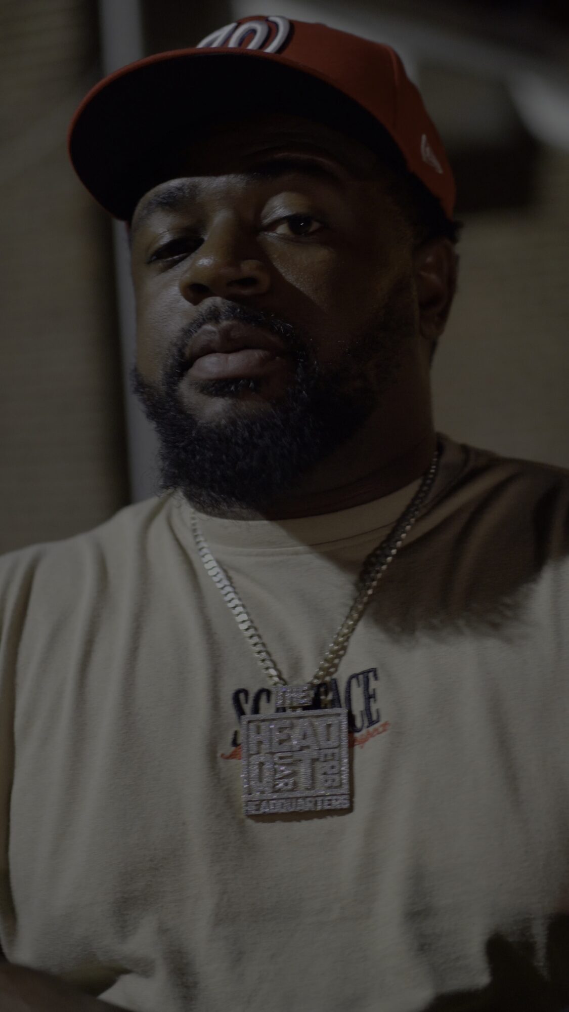 Man wearing a red cap, beige shirt, and a chain with a pendant, looking at the camera.