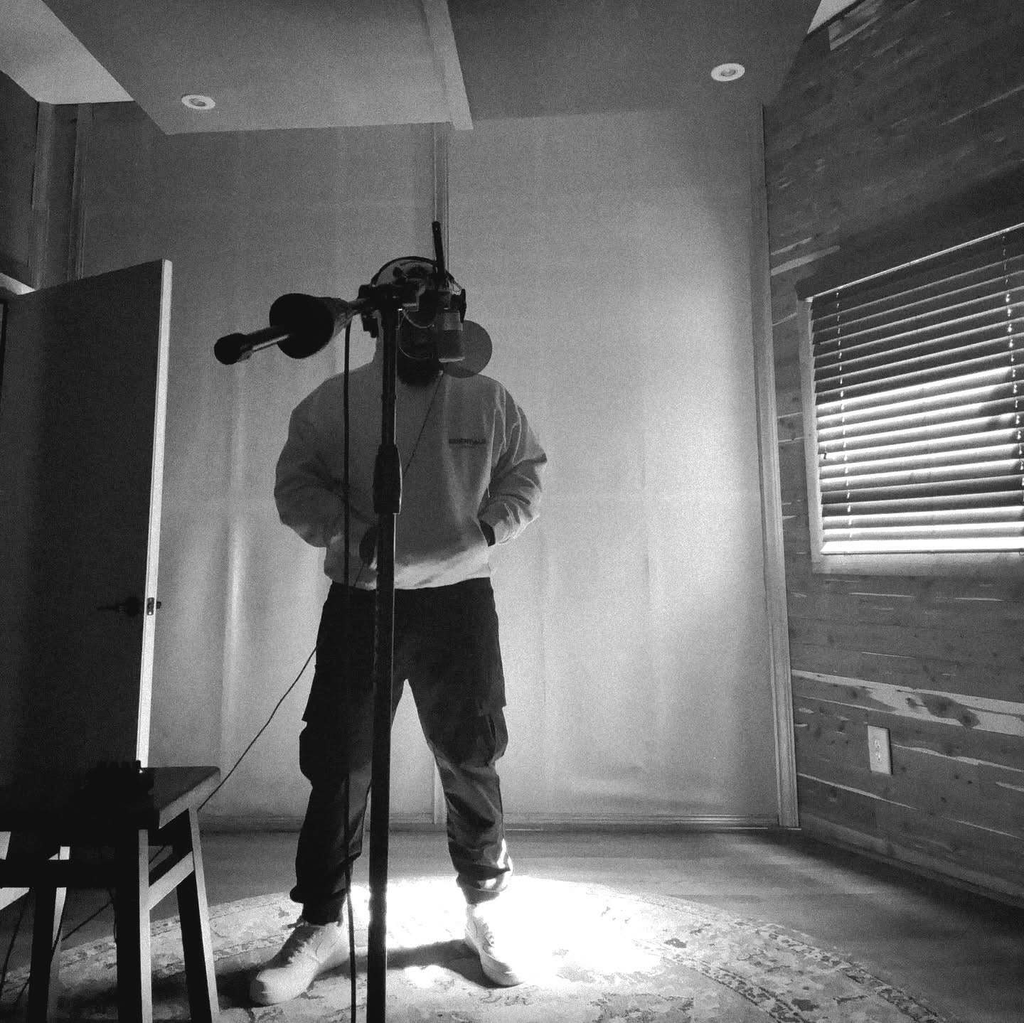 Person standing in front of a microphone in a room with wooden walls and a window with blinds.