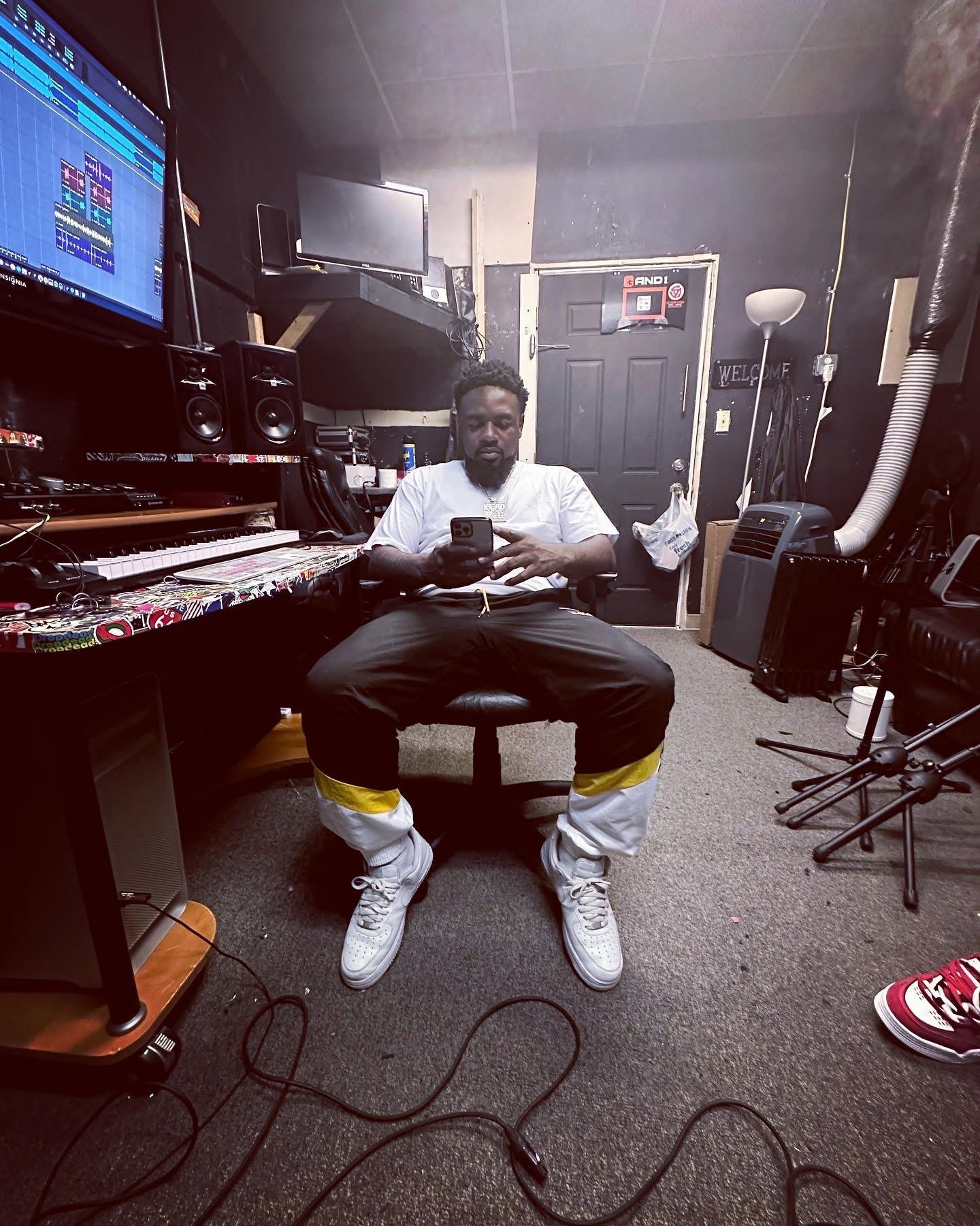 Person sitting in a music studio, holding a phone, surrounded by equipment and instruments, with a door in the background.