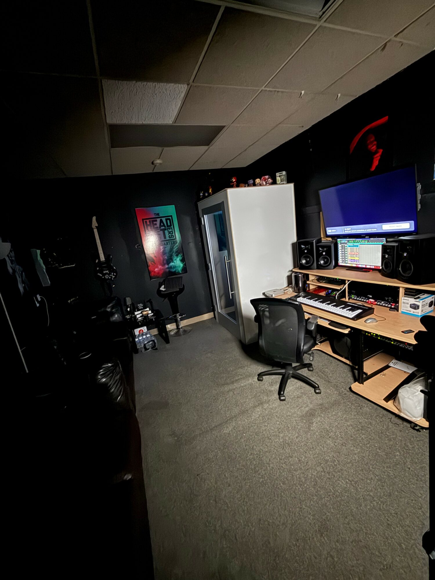 Recording studio with computer, speakers, and equipment, dimly lit room with a black ceiling and carpeted floor.