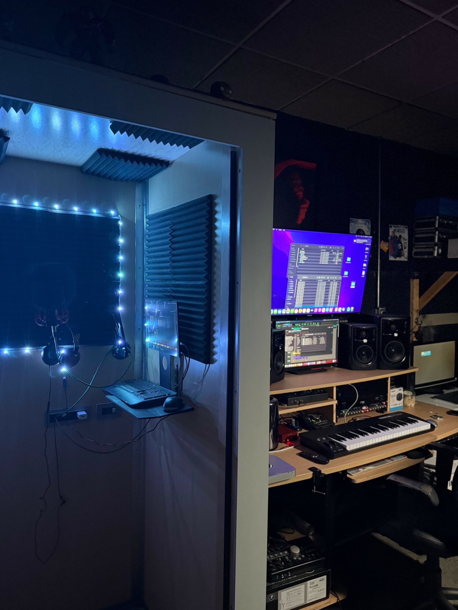 Music studio with a small vocal booth illuminated by blue LED lights, and a computer workstation with audio equipment.