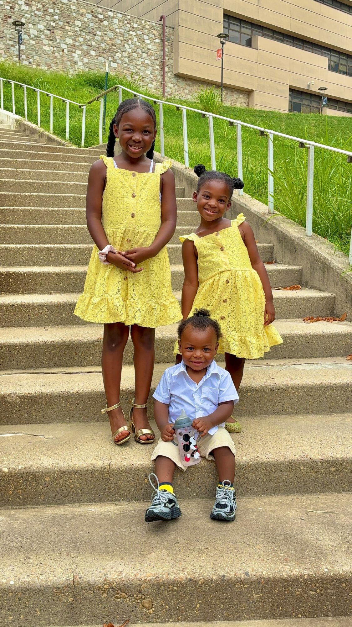 Three children, two girls and one boy, sitting and standing on outdoor concrete stairs with a railing and building in background.