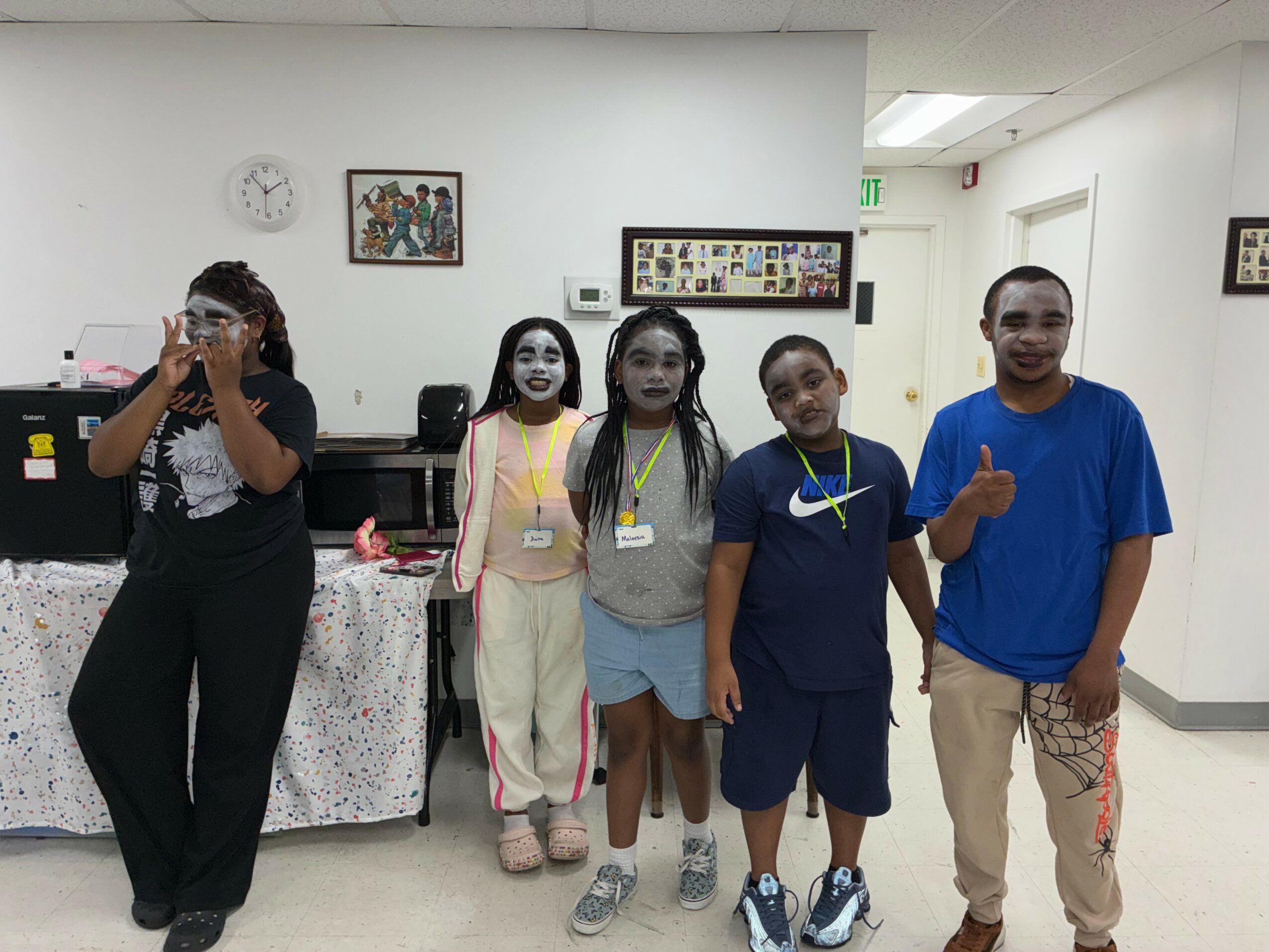 Five people standing in a room, some wearing face masks, with framed pictures on the wall behind them.