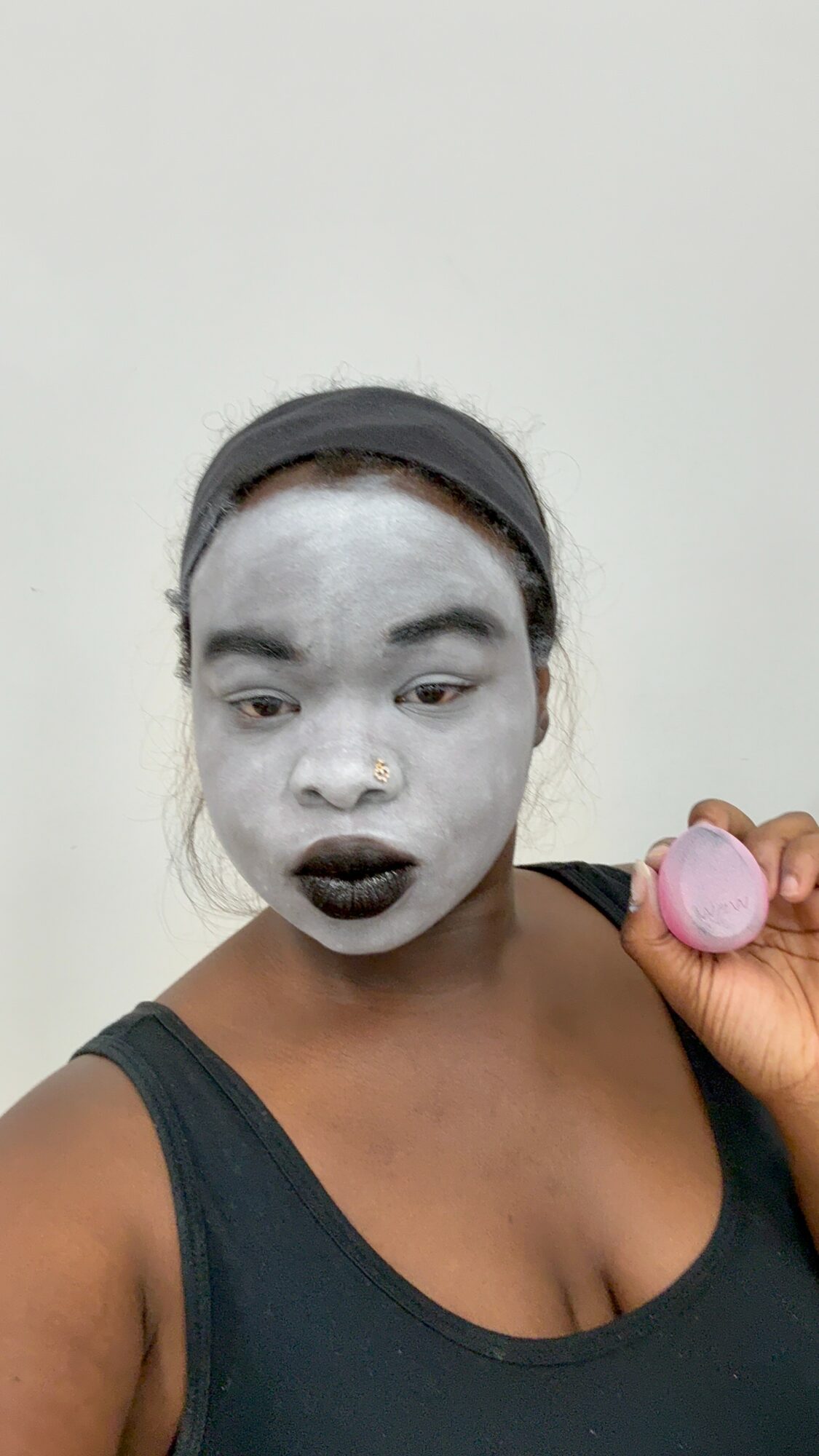Person with face painted white, dark lipstick, holding pink object, wearing black tank top, against plain background.