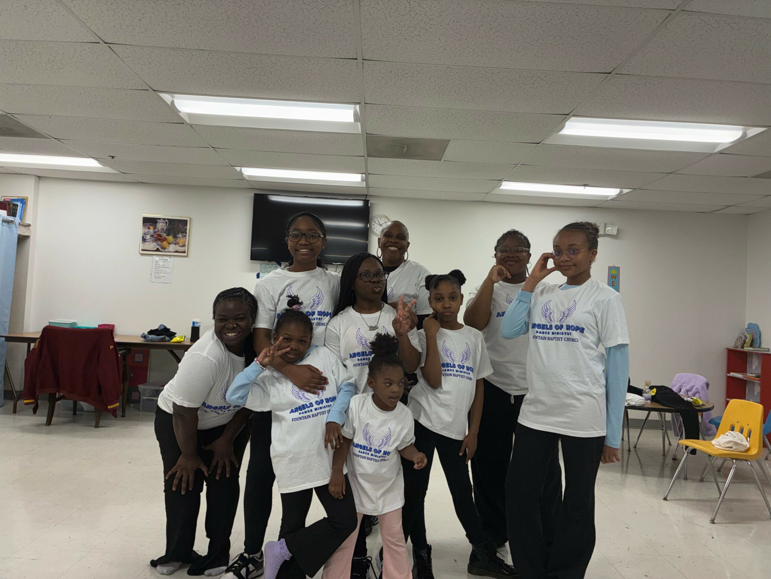 Group of children and adults in a room, some wearing matching white shirts, smiling and posing for the photo.