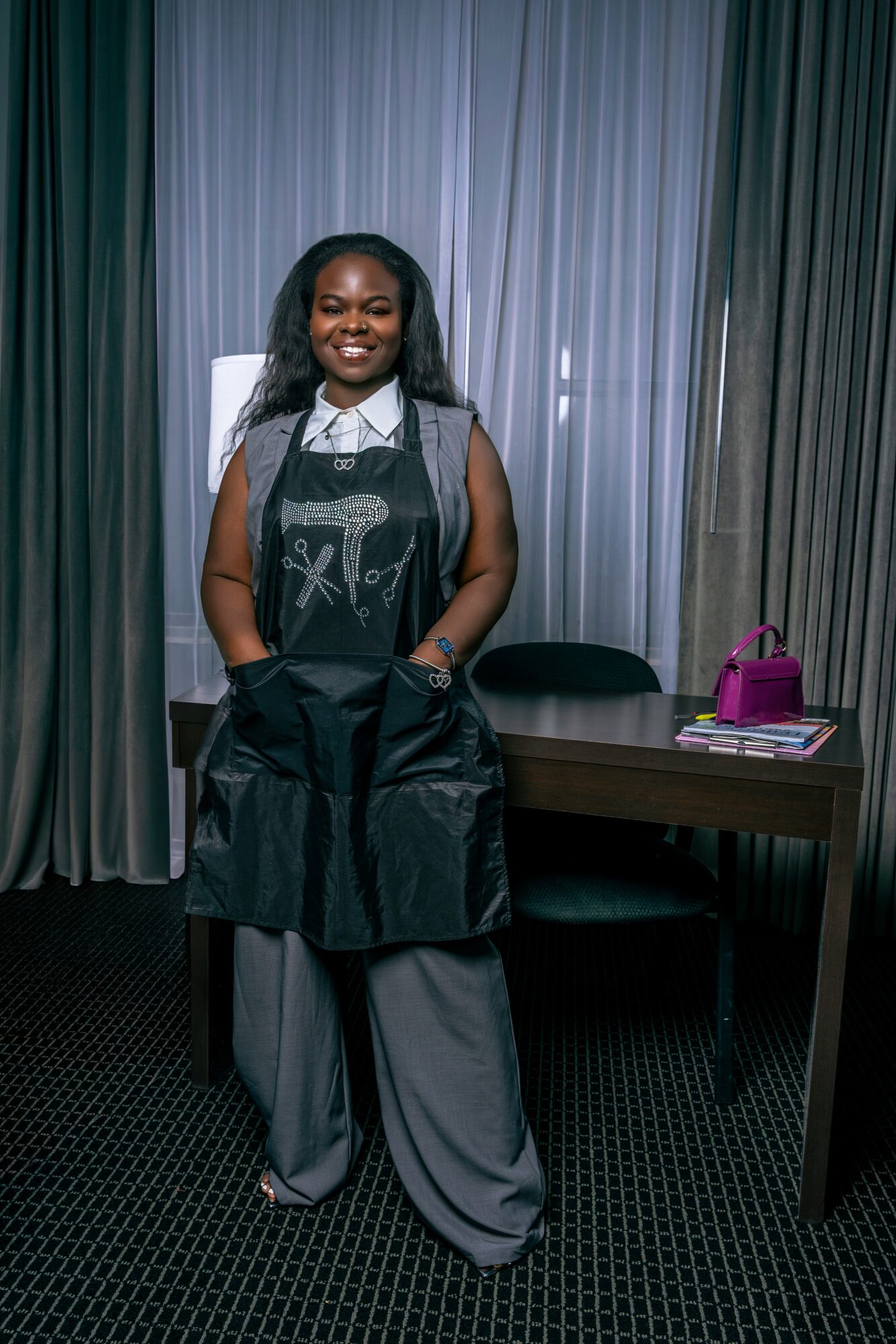 Smiling woman standing in front of curtains, wearing a black apron over gray pants, with a purple bag on desk nearby.