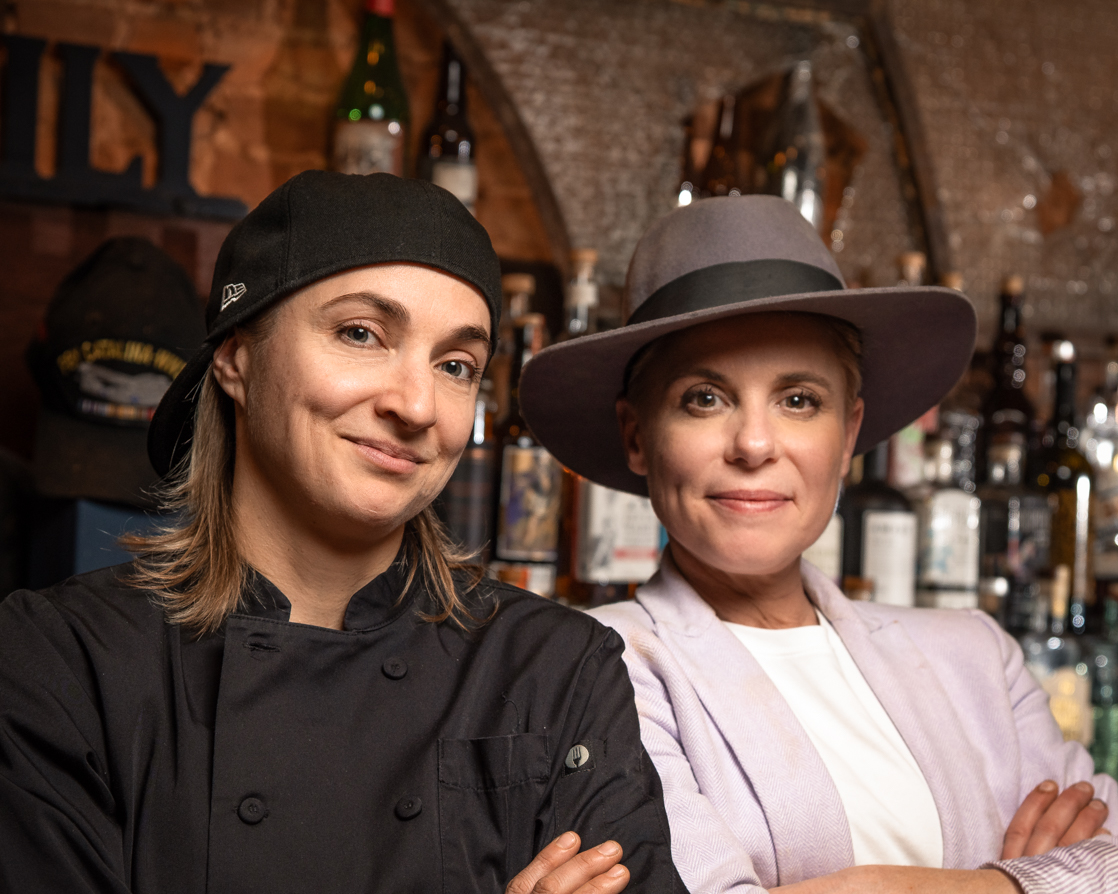 Two women stand side by side in a bar, one in a chef's uniform and the other in a hat and blazer, smiling.