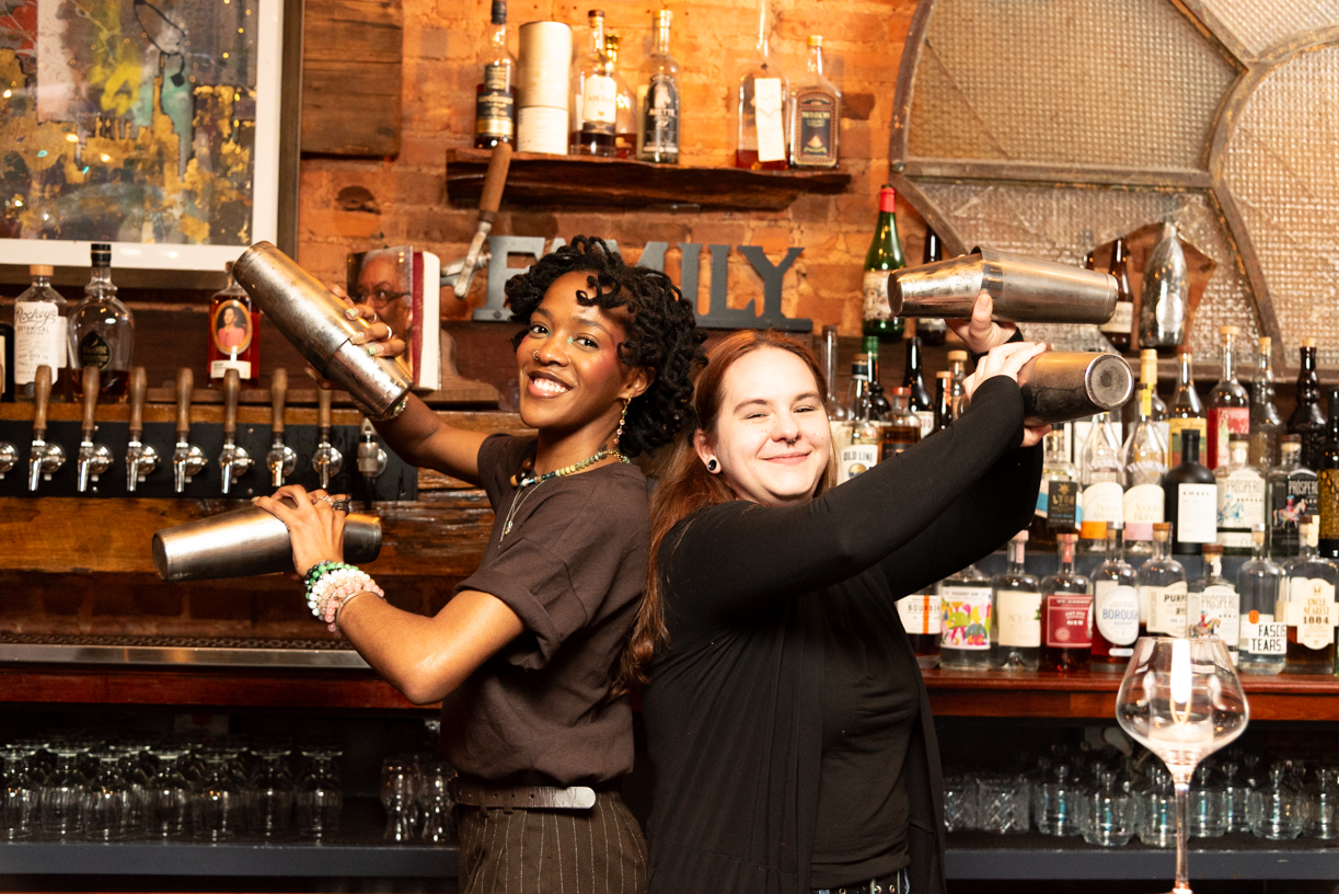 Two women behind a bar, smiling and holding beer taps, with bottles and glasses in the background.